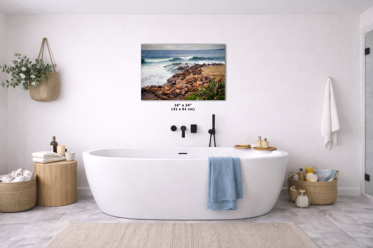 A coastal landscape print hangs above a white freestanding bathtub in a bright, modern bathroom. The artwork shows ocean waves, rocks, and a sandy shore.