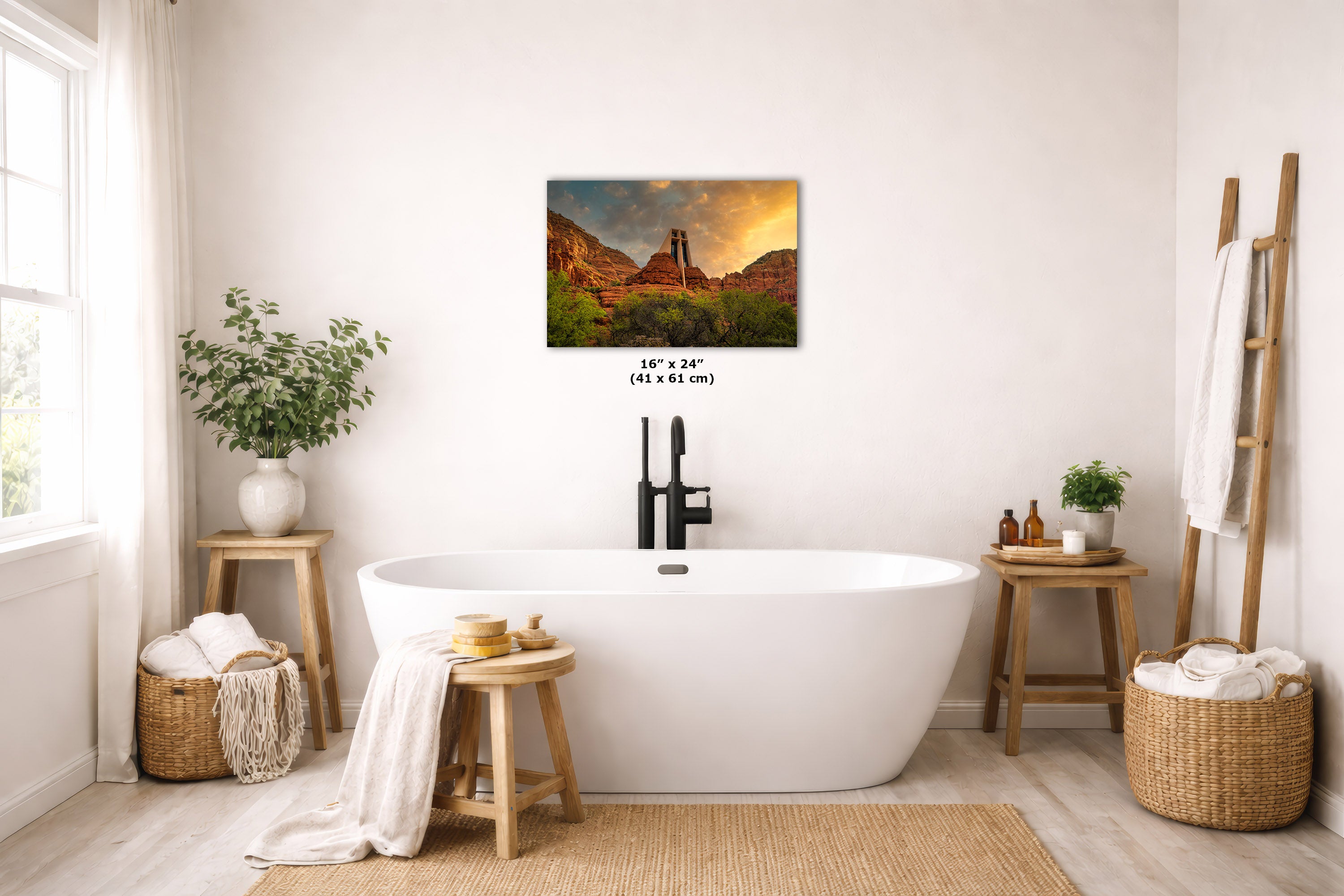 A wall art print depicting a mountain landscape with a pink and orange sunset sky, displayed above a white freestanding bathtub in a bright, modern bathroom with wooden stools, plants, and woven baskets.