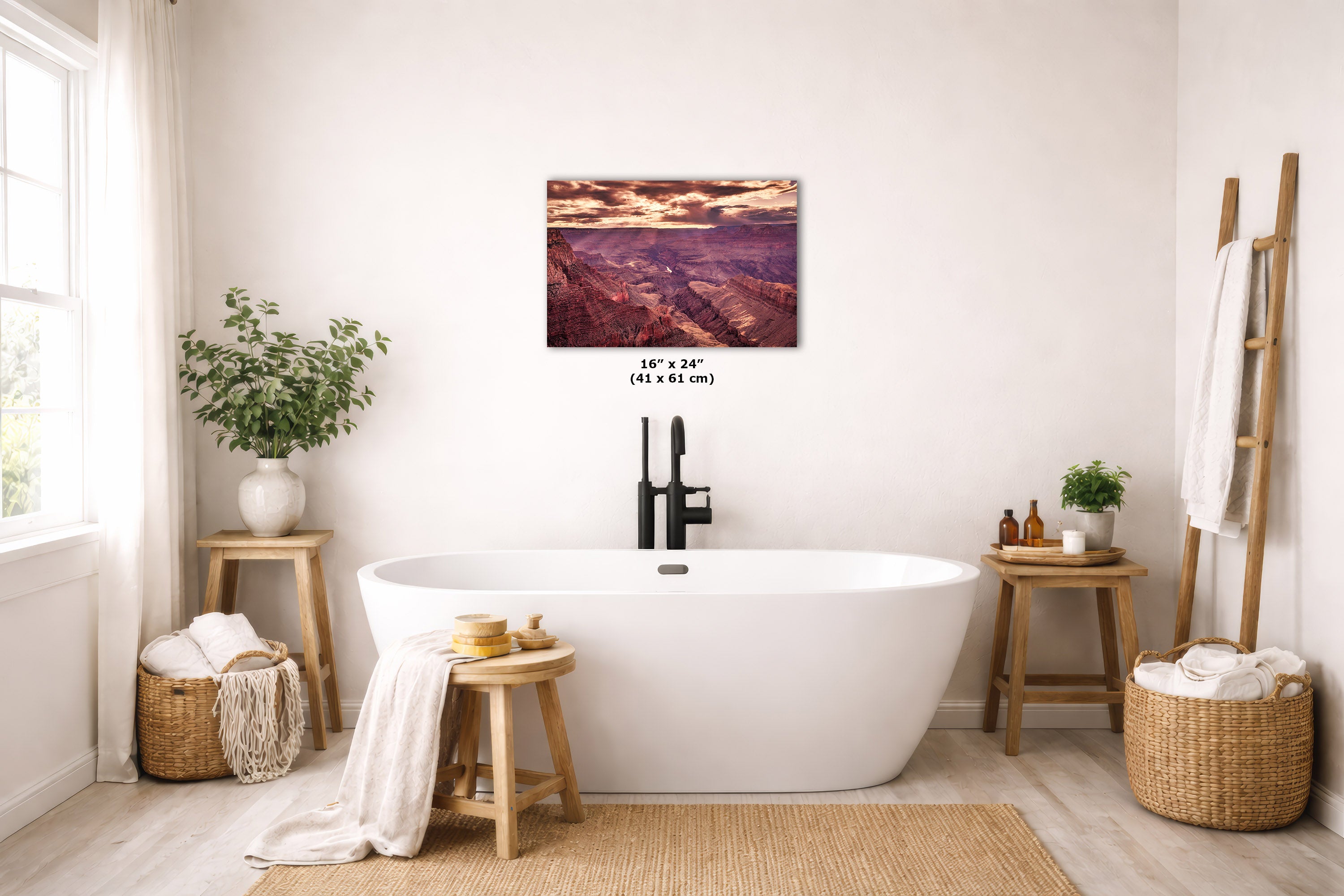 A wall art print depicting a mountain landscape with a pink and orange sunset sky, displayed above a white freestanding bathtub in a bright, modern bathroom with wooden stools, plants, and woven baskets.