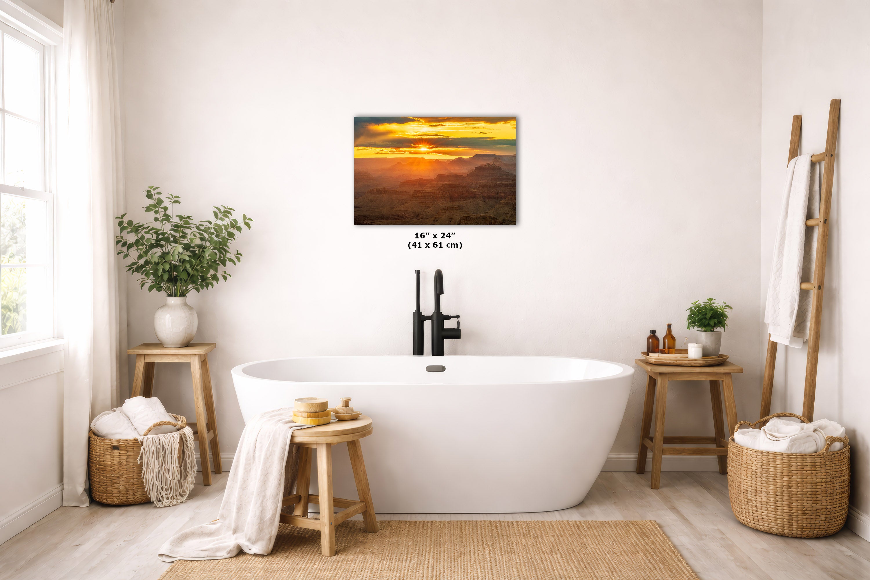 A wall art print showing a warm sunset over a mountain landscape, displayed above a white freestanding bathtub in a bright, neutral-toned bathroom with wooden stools, plants, baskets, and a ladder shelf.