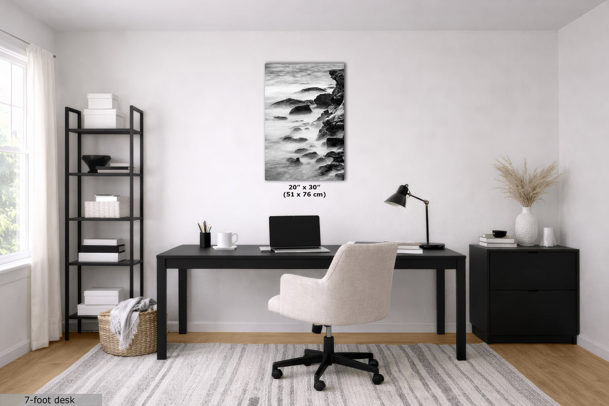A modern home office setup featuring a black desk with a light beige swivel chair, a black shelving unit, and a black side cabinet. The focal point is a black and white vertical wall art print depicting a rocky shoreline, with dimensions '20" x 30"' printed below it.