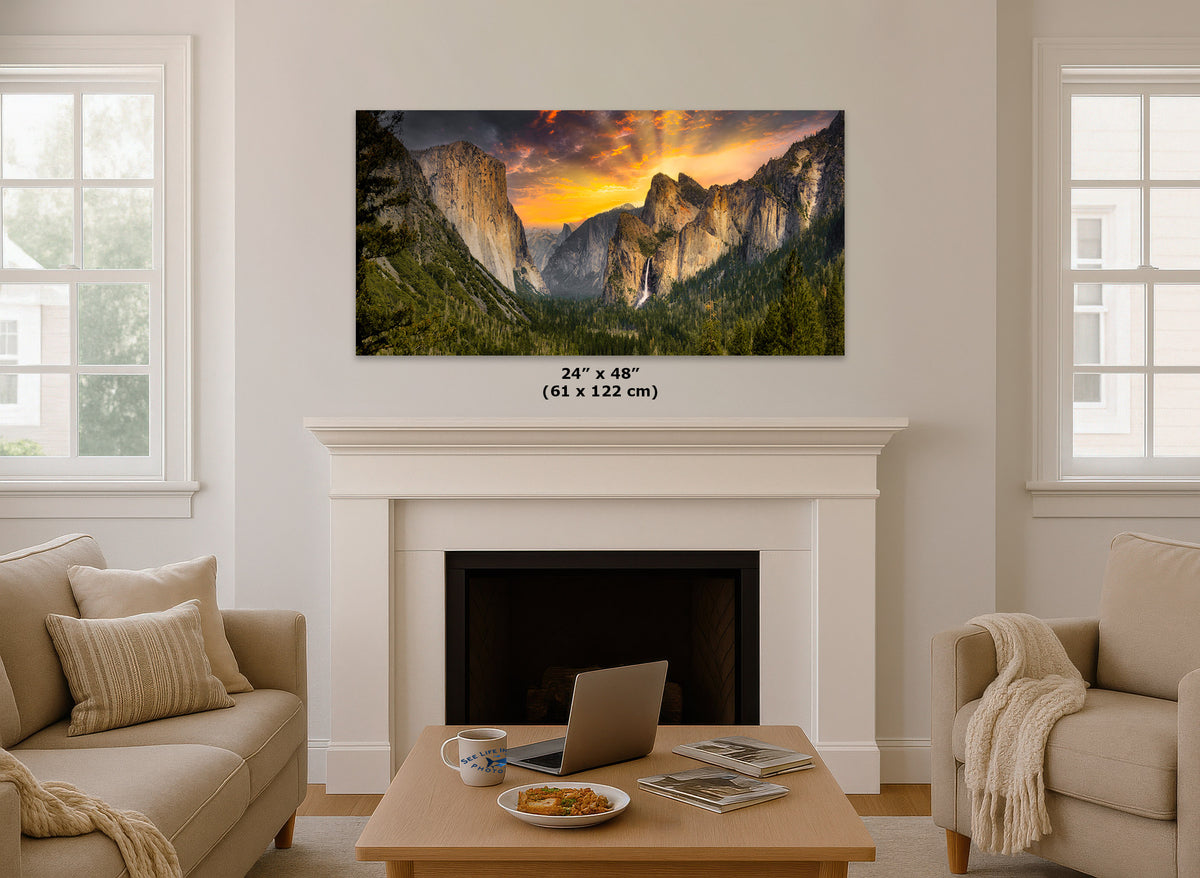 A Yosemite Tunnel View wall art print of a mountain valley landscape with a dramatic orange sunset is displayed above a white fireplace mantel in a bright living room. The print includes size text reading 24 x 4 inches (61 x 122 cm).