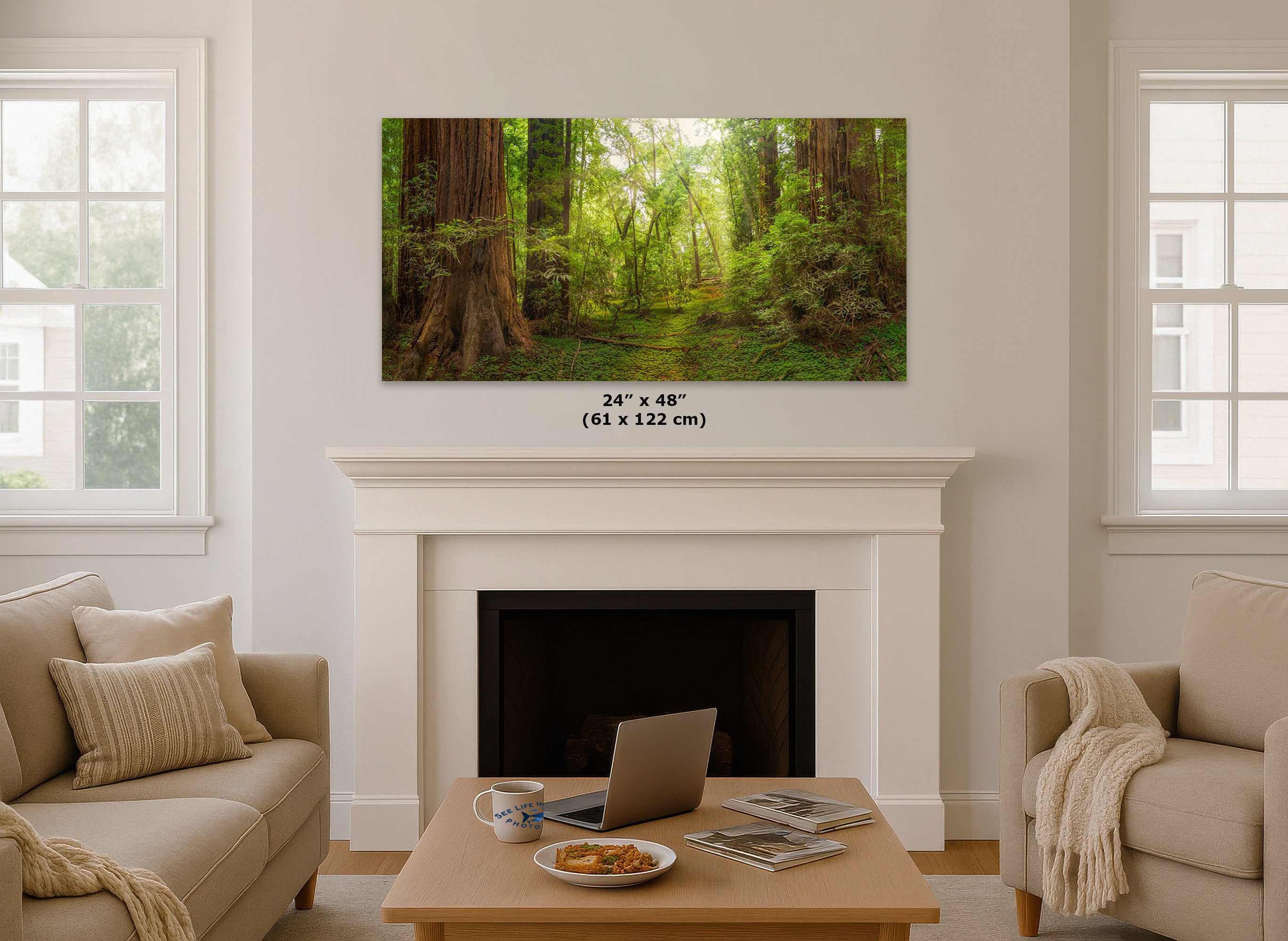 A framed forest wall art print showing a lush green woodland path with tall trees, displayed above a white fireplace in a bright living room with beige sofas and a coffee table.