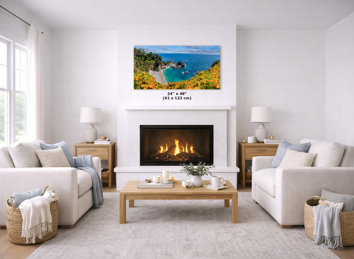 A framed coastal landscape wall art print hangs above a fireplace in a bright, modern living room. The print shows a sunny seaside cliff with blue water, green trees, and yellow flowers.