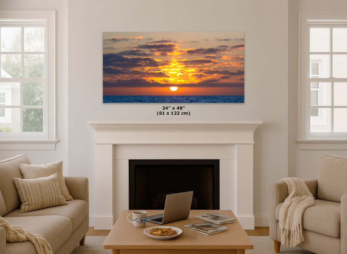 A 24x48 inch canvas print of a vibrant sunset over the ocean, displayed on a white fireplace mantel in a modern living room setting. The canvas features a warm orange and yellow sunset with dark blue water below.