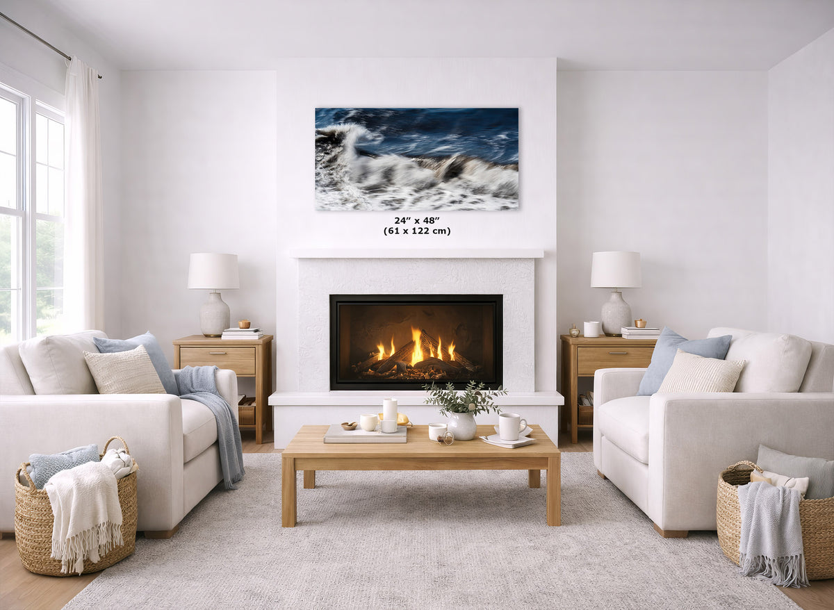 A framed wall art print depicting a dramatic ocean wave scene in blue, white, and gray tones, displayed above a fireplace in a bright, modern living room. The print includes a size label reading 24" x 48" (61 x 122 cm).