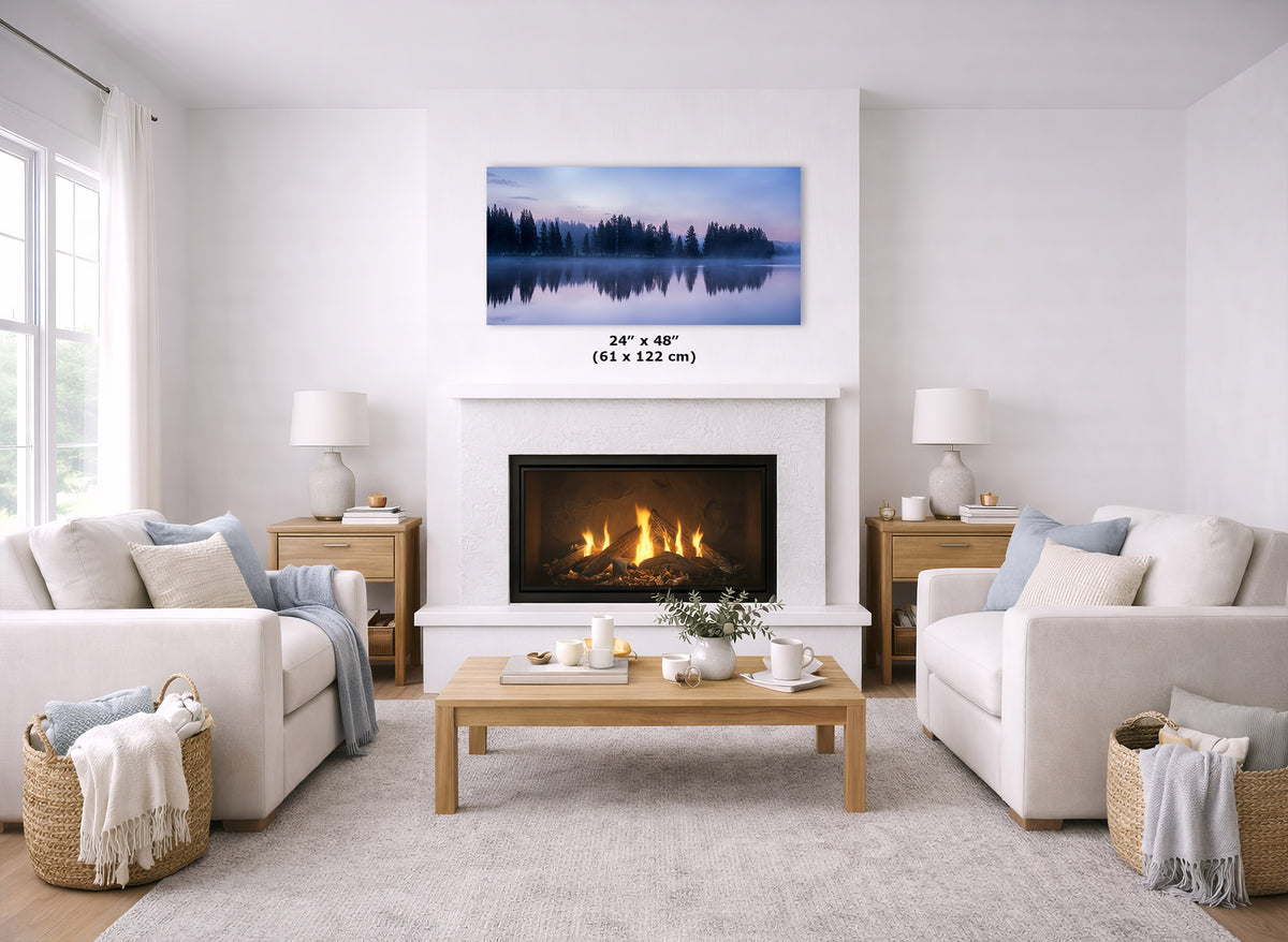 A framed landscape wall art print showing a calm lake and pine trees at dusk, displayed above a fireplace in a bright, modern living room. The print has a size label overlaid in the center reading 24" x 48" (61 x 122 cm).