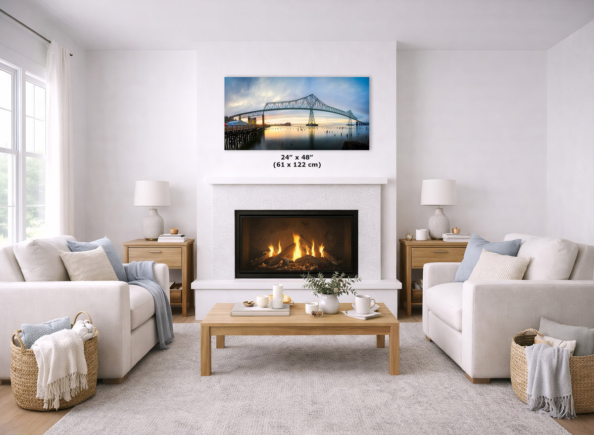 A framed wall art print of a bridge over water is displayed above a fireplace in a bright, modern living room with two white sofas, a wooden coffee table, and a light rug. The print includes a size measurement label reading 24" x 48" (61 x 122 cm).