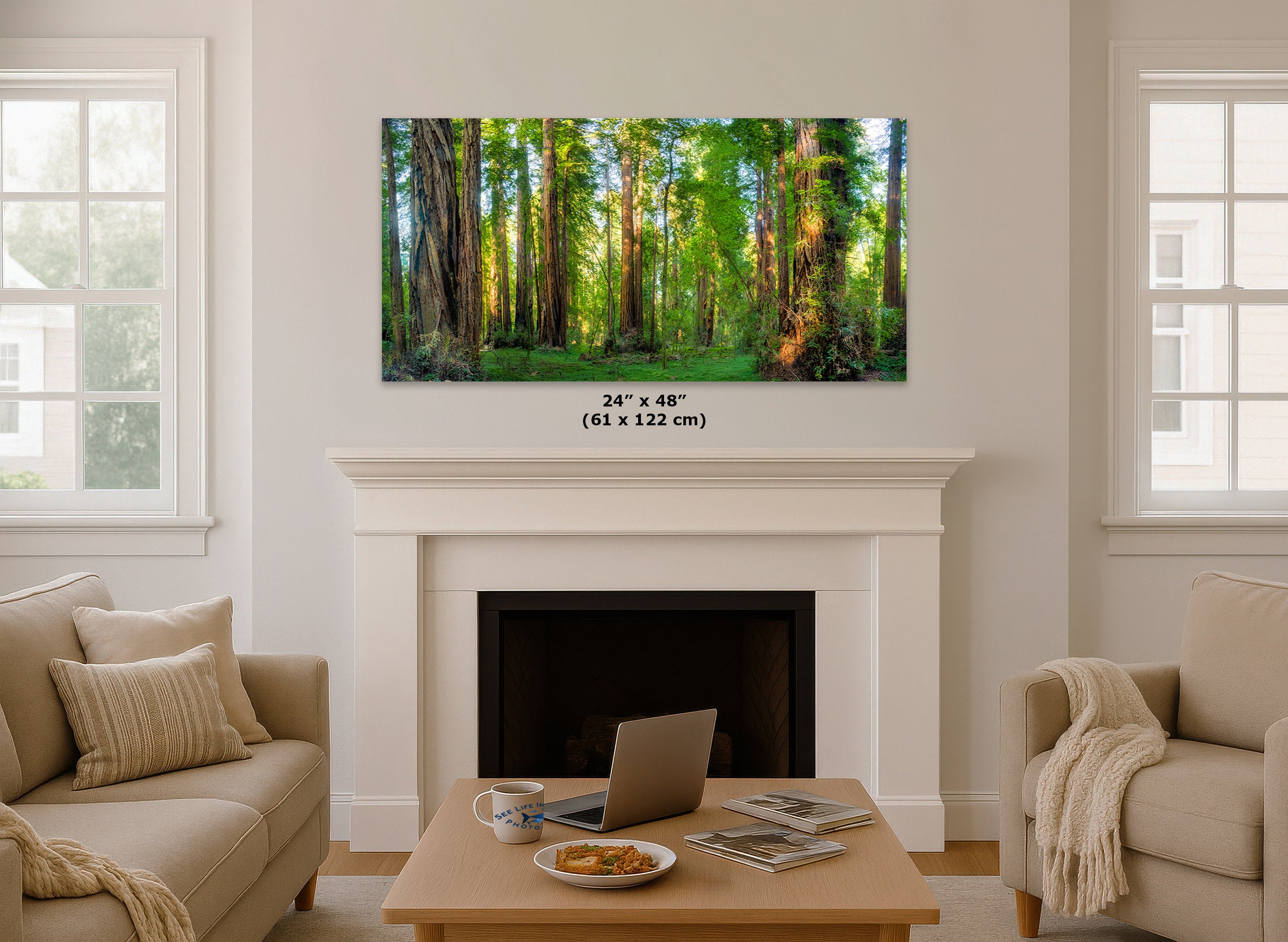 A framed forest wall art print hangs above a white fireplace mantel in a bright living room with beige sofas, a coffee table, and windows on both sides. The print depicts tall green trees with warm sunlight filtering through the canopy.