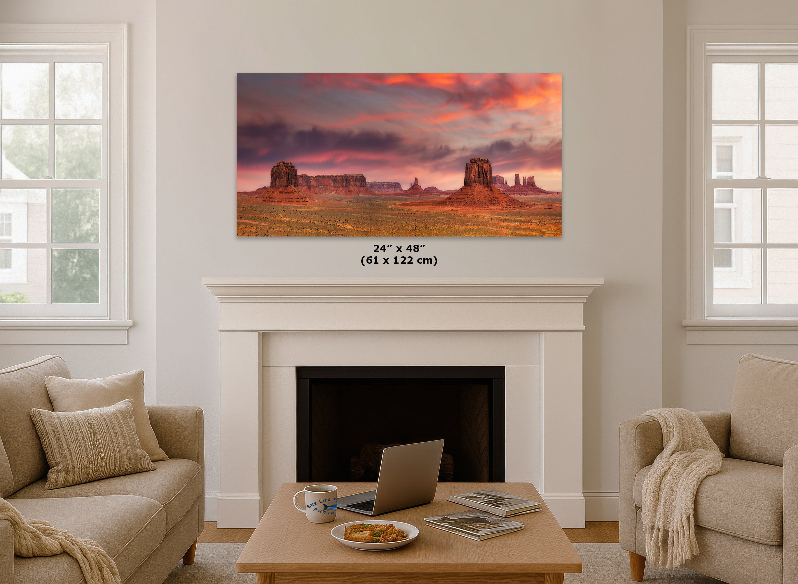 A wall art print showing a desert landscape with rock formations under a dramatic pink and orange sky, displayed above a white fireplace in a bright living room. The print includes size text reading 24 x 48 inches (61 x 122 cm).