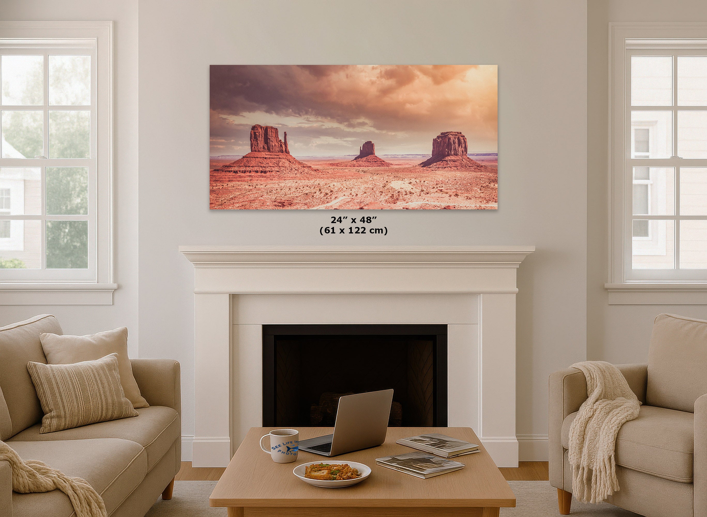 A wall art print showing a desert landscape with red rock buttes under a dramatic sky, displayed above a white fireplace in a living room. The print includes size text reading 24 x 48 inches (61 x 122 cm).