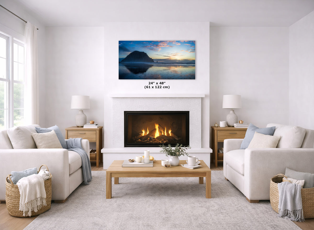 A landscape wall art print showing a sunset over water with a dark island silhouette, displayed above a fireplace in a bright, modern living room. The print includes a size overlay reading 24" x 48" (61 x 122 cm).