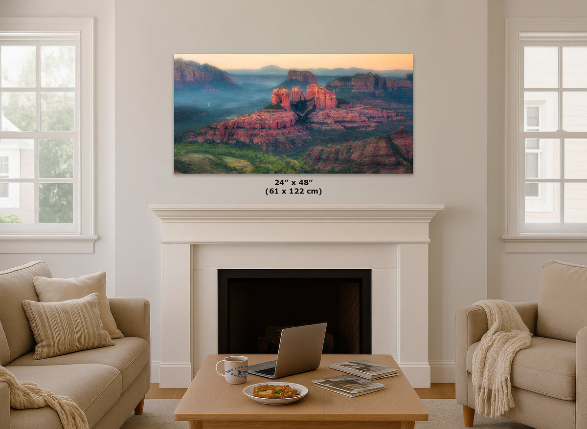 A wall art print showing a desert canyon landscape with red rock formations at sunset, displayed above a white fireplace in a bright living room. The print includes size text reading 24 x 48 inches (61 x 122 cm).