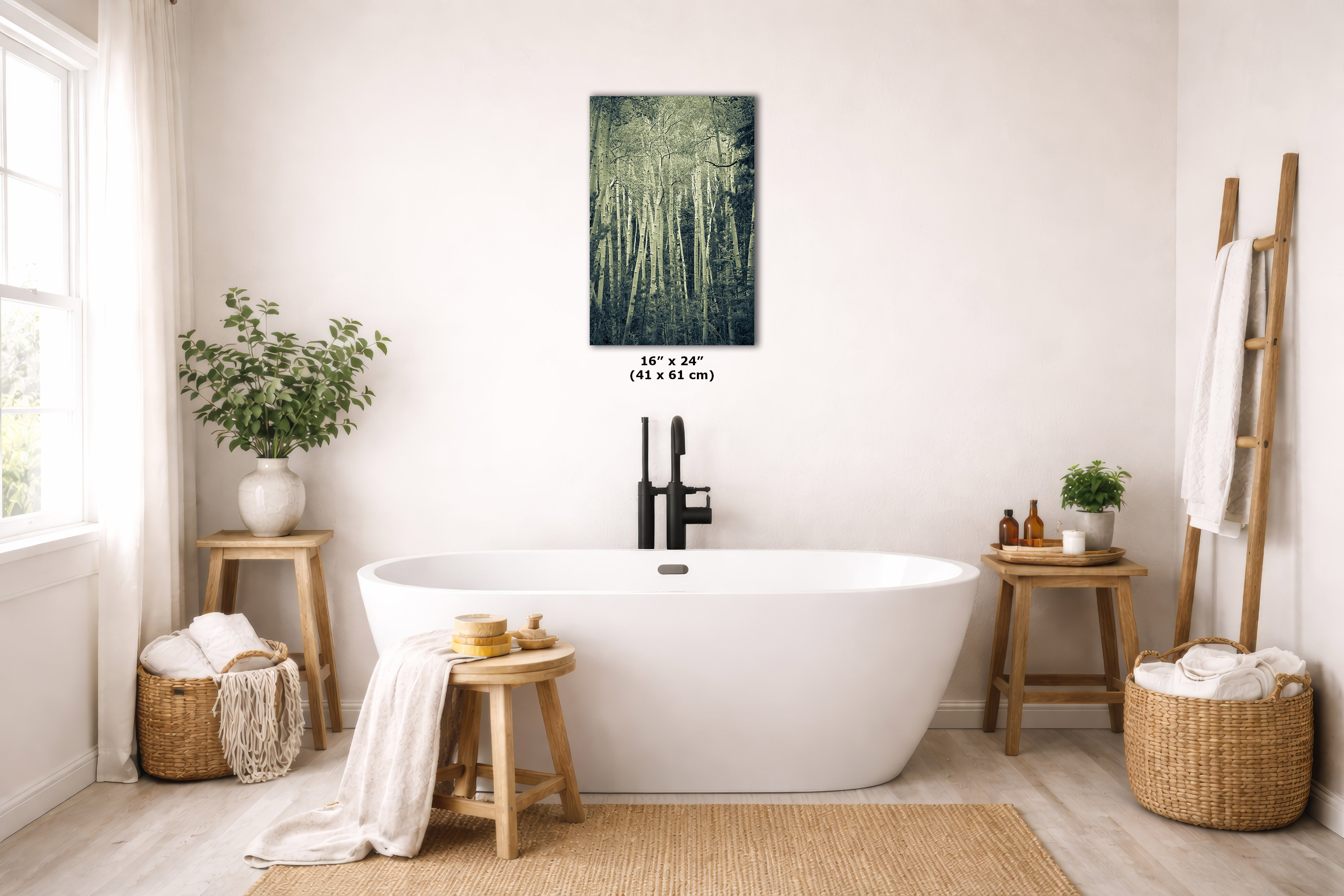A freestanding white bathtub in a bright, neutral-toned bathroom with a wall-mounted vertical print of a green forest scene above it. The print includes size text reading 16 x 24 inches.