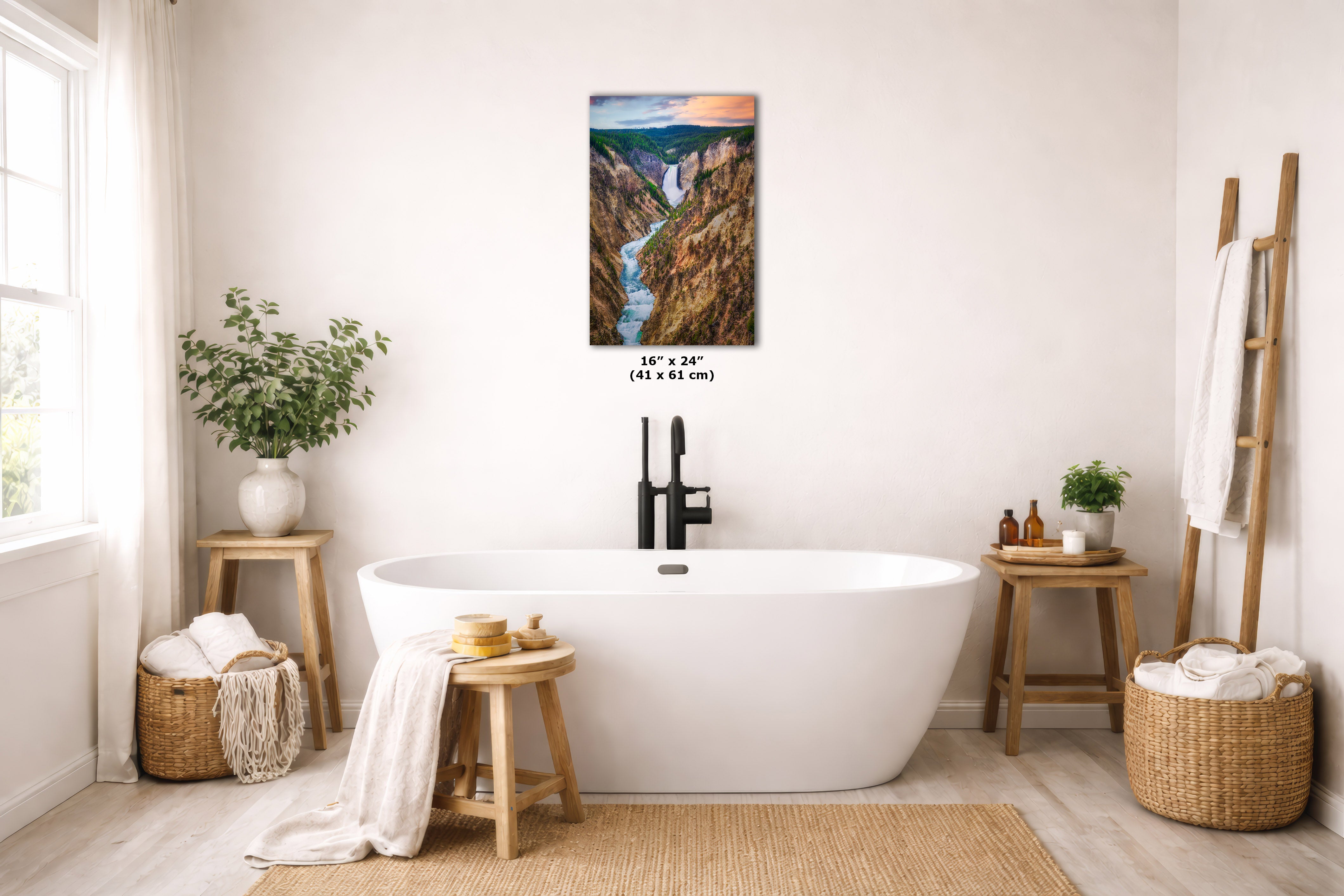 A framed vertical landscape art print depicting a canyon river scene, displayed on a light beige wall above a white freestanding bathtub in a bright, modern bathroom setting.