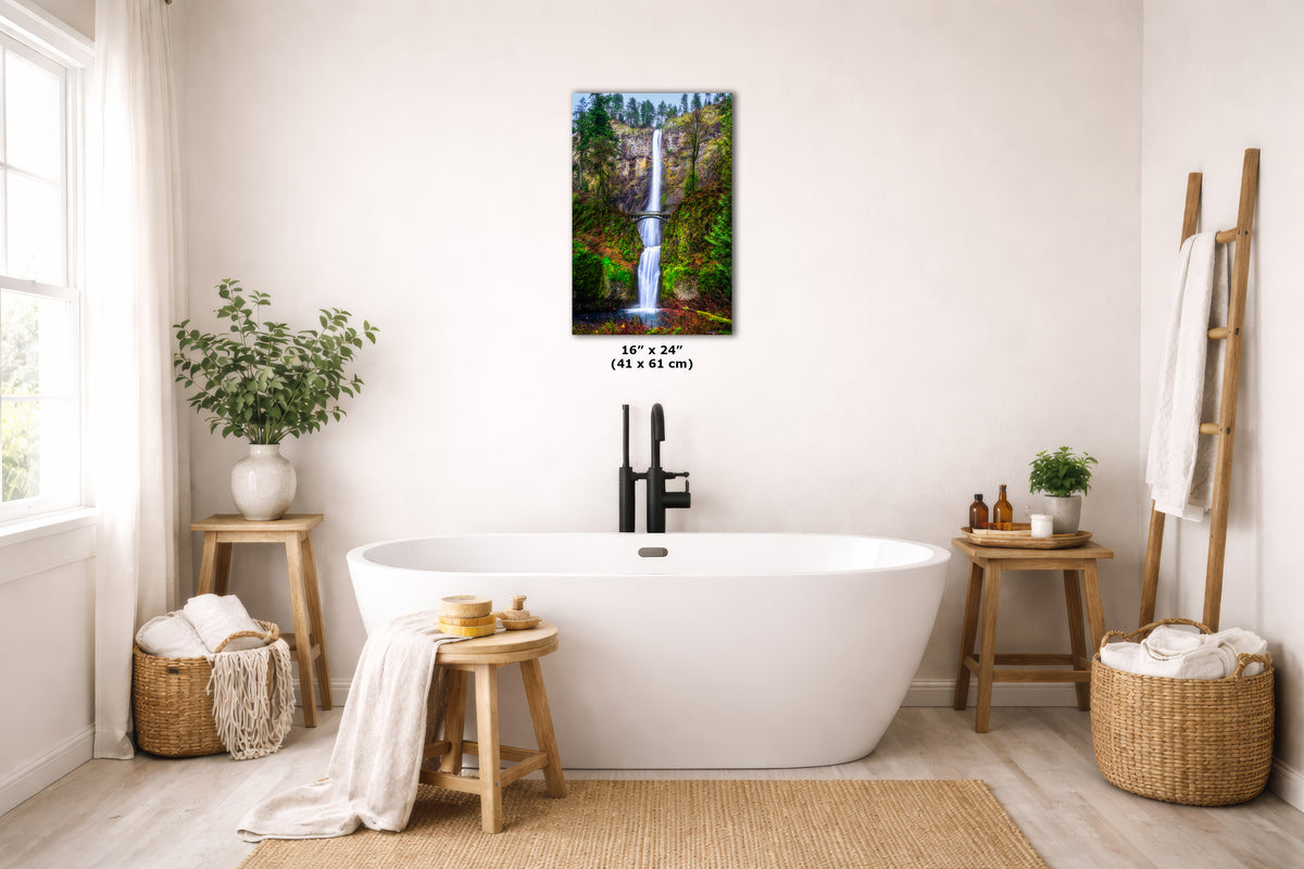 A framed wall art print depicting a waterfall in a forest, displayed above a white freestanding bathtub in a bright, modern bathroom setting.
