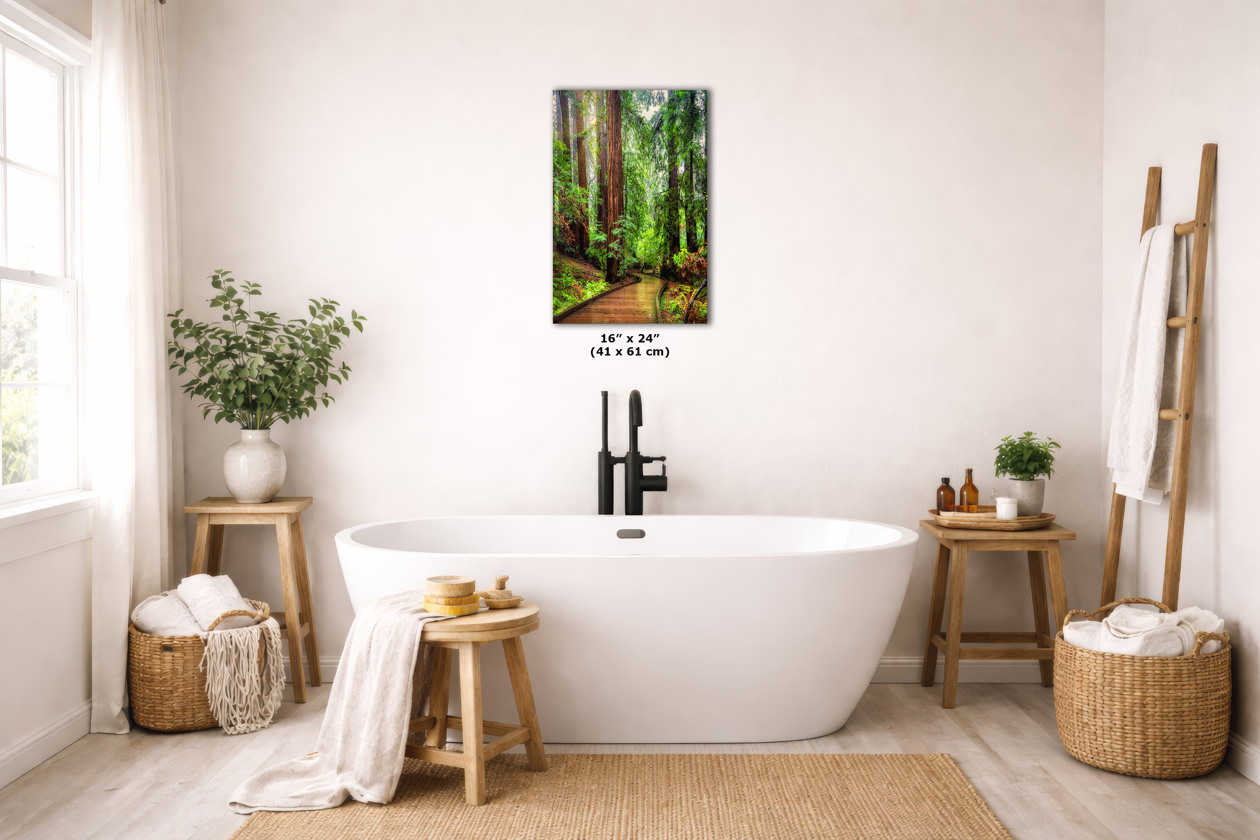 A framed forest wall art print hangs above a white freestanding bathtub in a bright, neutral bathroom. The artwork shows a sunlit forest path with tall trees and lush green foliage.
