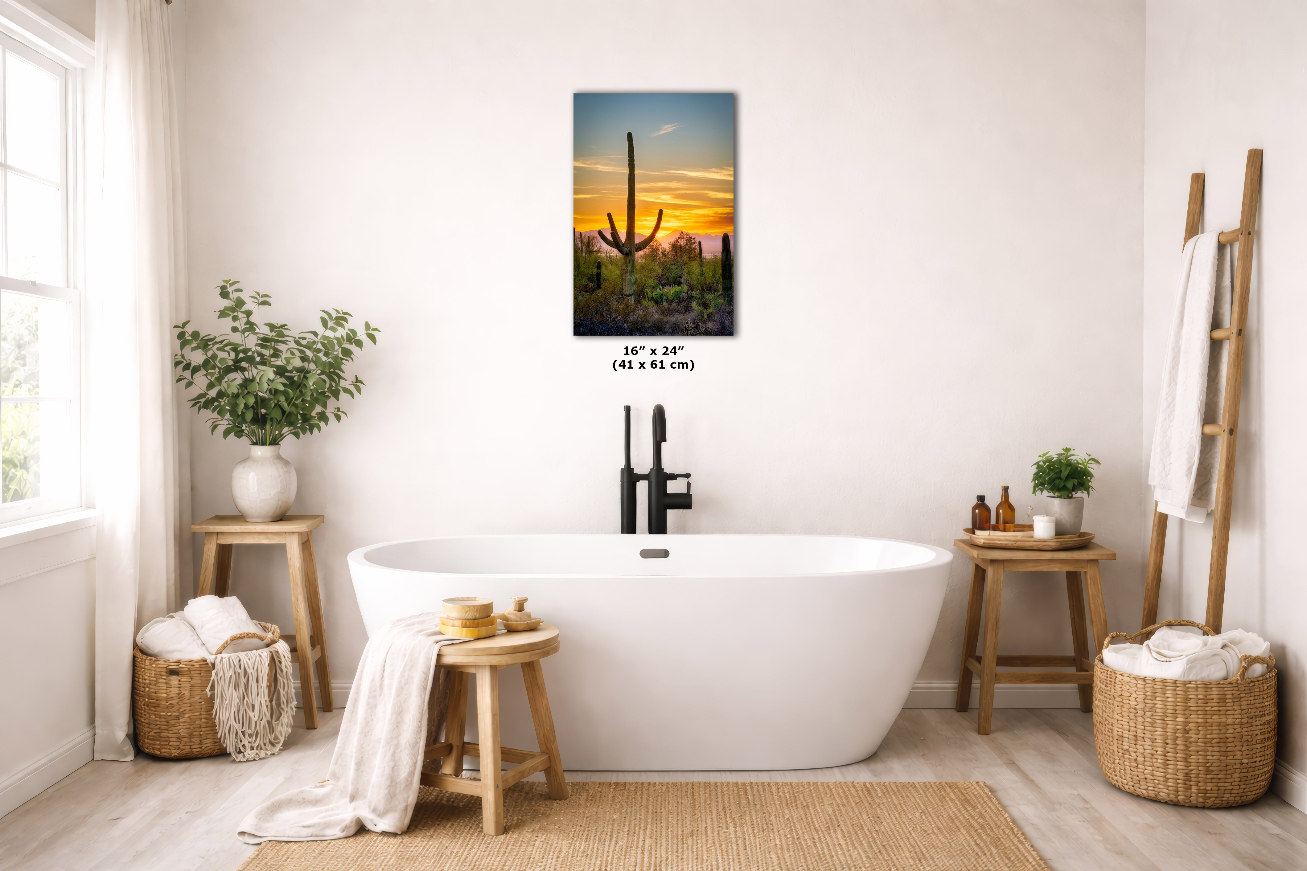 A framed vertical canvas print of a desert sunset with saguaro cacti hangs on a light beige wall above a white freestanding bathtub in a bright, modern bathroom. The print includes size text reading 16 x 24 in (41 x 61 cm) below it.