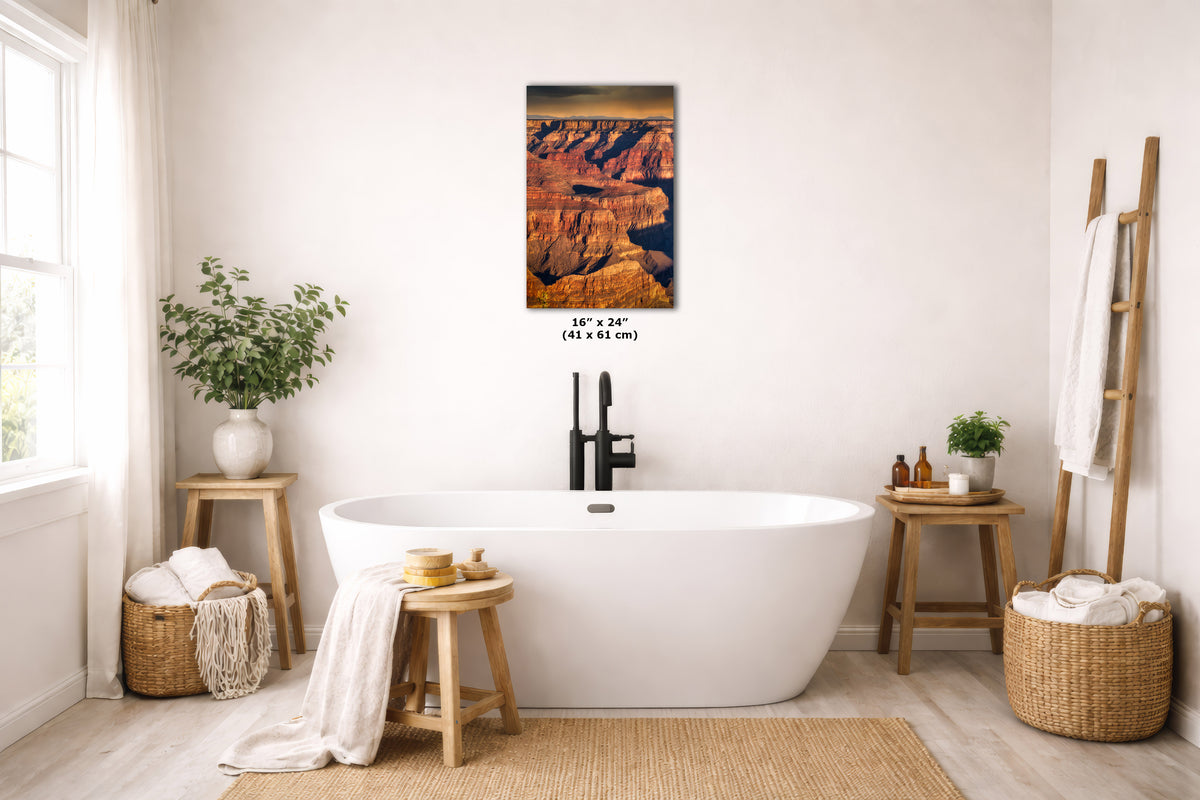 A rectangular wall art print depicting a canyon landscape in warm orange, brown, and black tones, displayed above a white freestanding bathtub in a bright, neutral bathroom with wooden stools, baskets, and plants.