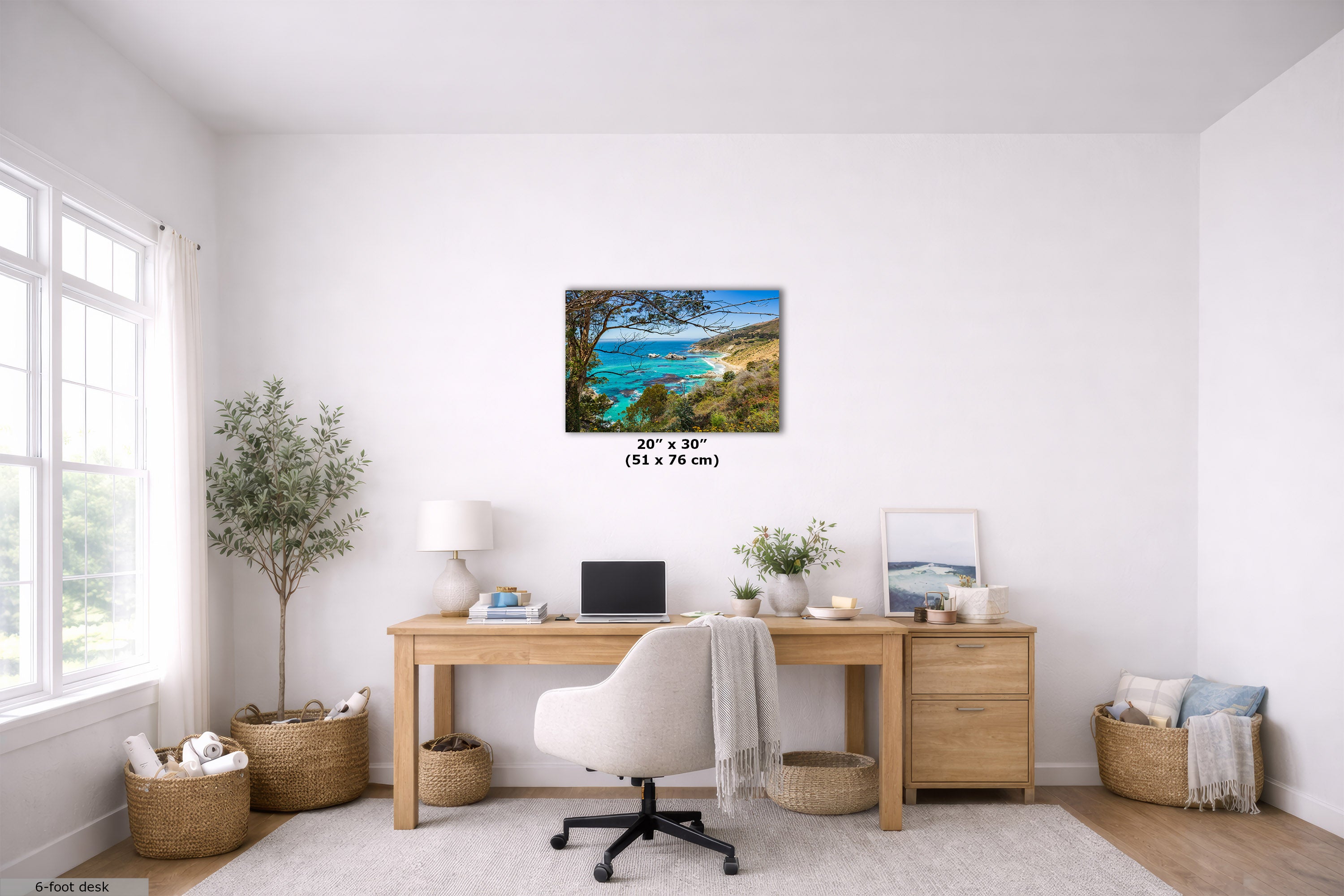 A 20" x 30" (51 cm x 76 cm) landscape wall art print of a Big Sur, California, coastal beach with turquoise water and green trees, displayed on a white wall in a bright, modern home office setting above a wooden desk.