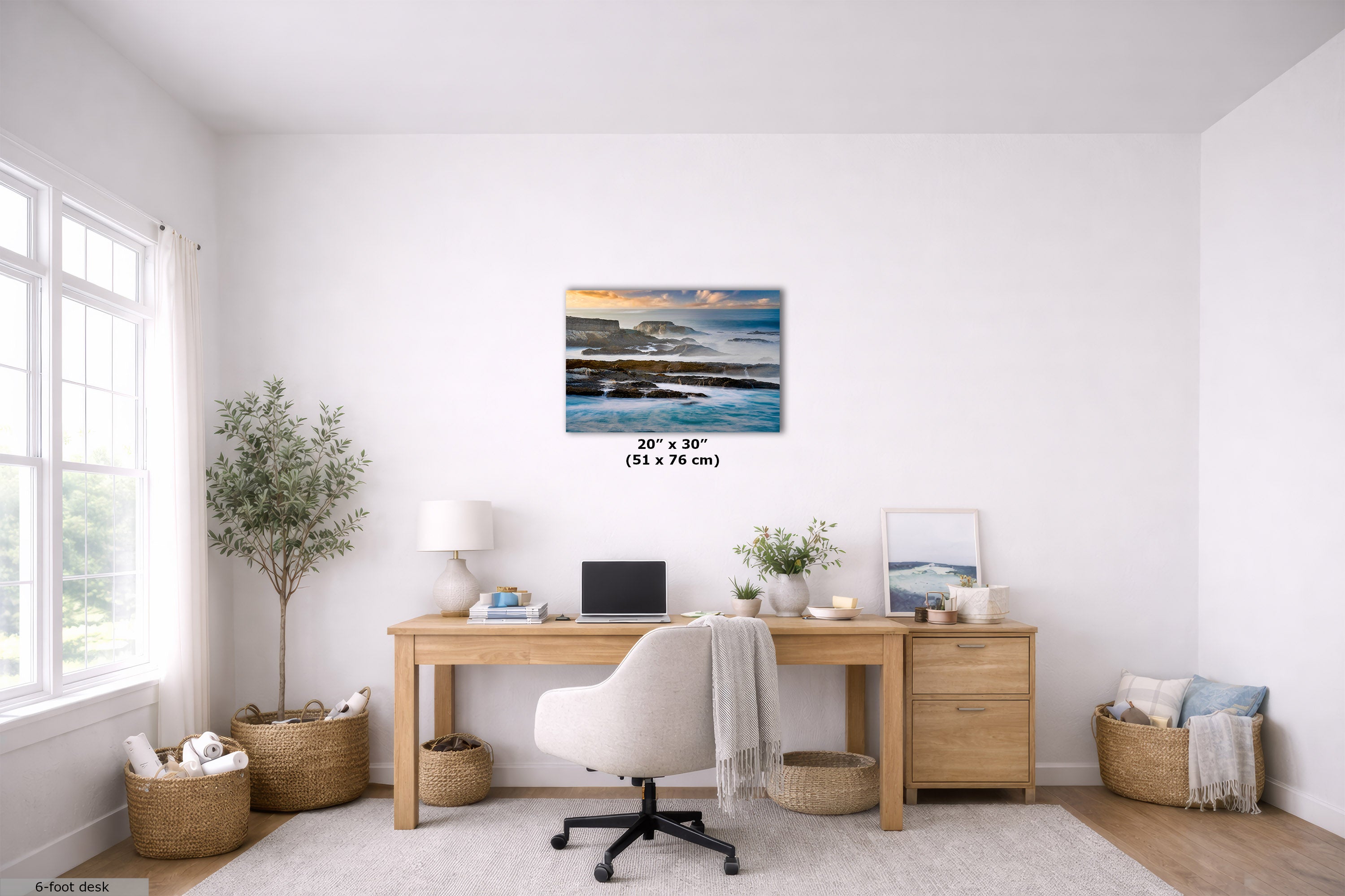 A 20" x 30" canvas print of a rocky beach sunset is displayed on a white wall in a bright, modern home office setting with a wooden desk, white chair, and plants.