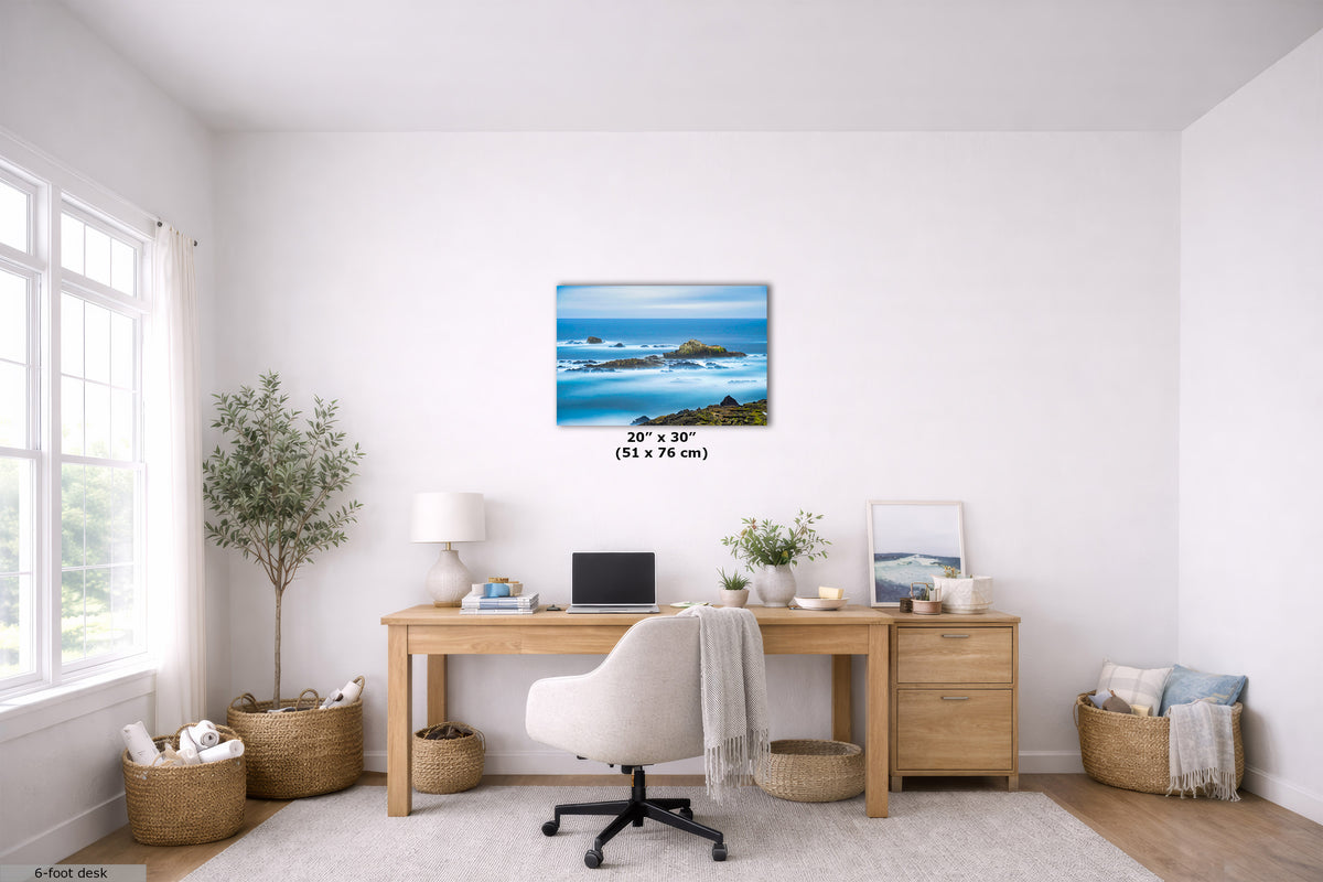 A 23 x 30 inch wall art print depicting a scenic blue ocean and rocky coastline, displayed in a bright, modern home office setting above a wooden desk.