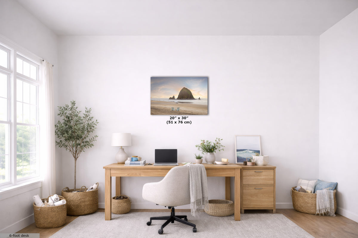 A 20" x 30" canvas print of Haystack Rock on a beach is displayed on a white wall in a bright, modern home office setting. The room features a light wood desk, a white office chair, potted plants, and woven baskets.