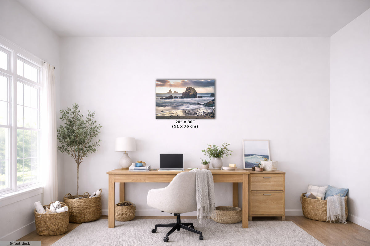 A 20" x 30" (51 cm x 76 cm) canvas print of a coastal rock formation at sunset, displayed on a white wall in a modern, bright home office setting above a wooden desk.
