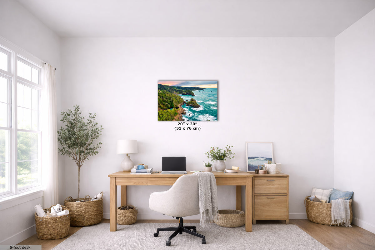 A coastal landscape wall art print hangs above a wooden desk in a bright, modern home office. The print depicts a rocky coastline with turquoise ocean waves and green cliffs under a sunset sky.