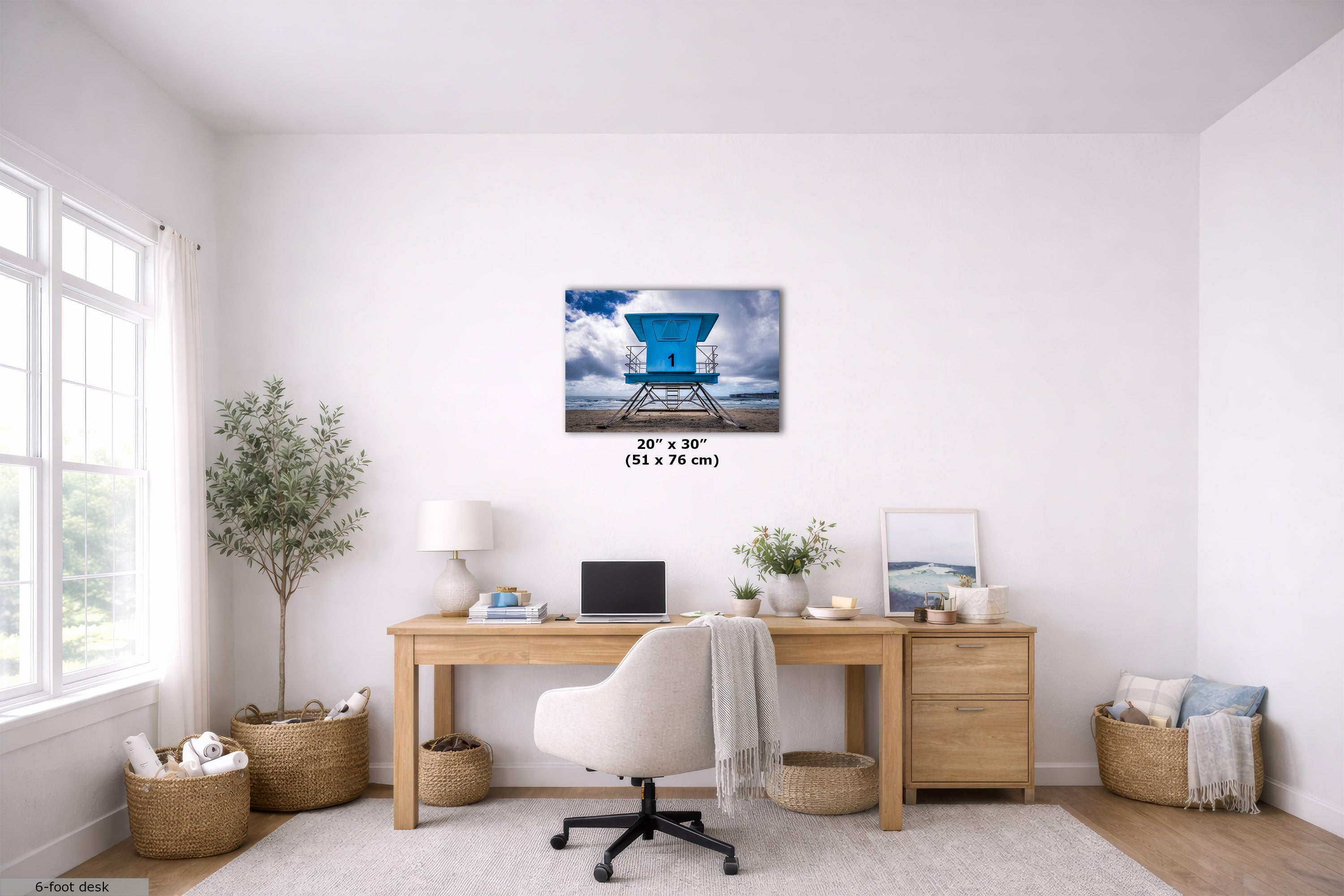 A wall art print showing a blue lifeguard tower over water, displayed above a wooden desk in a bright, modern home office. The print includes size text indicating 20" x 30" (51 x 76 cm).