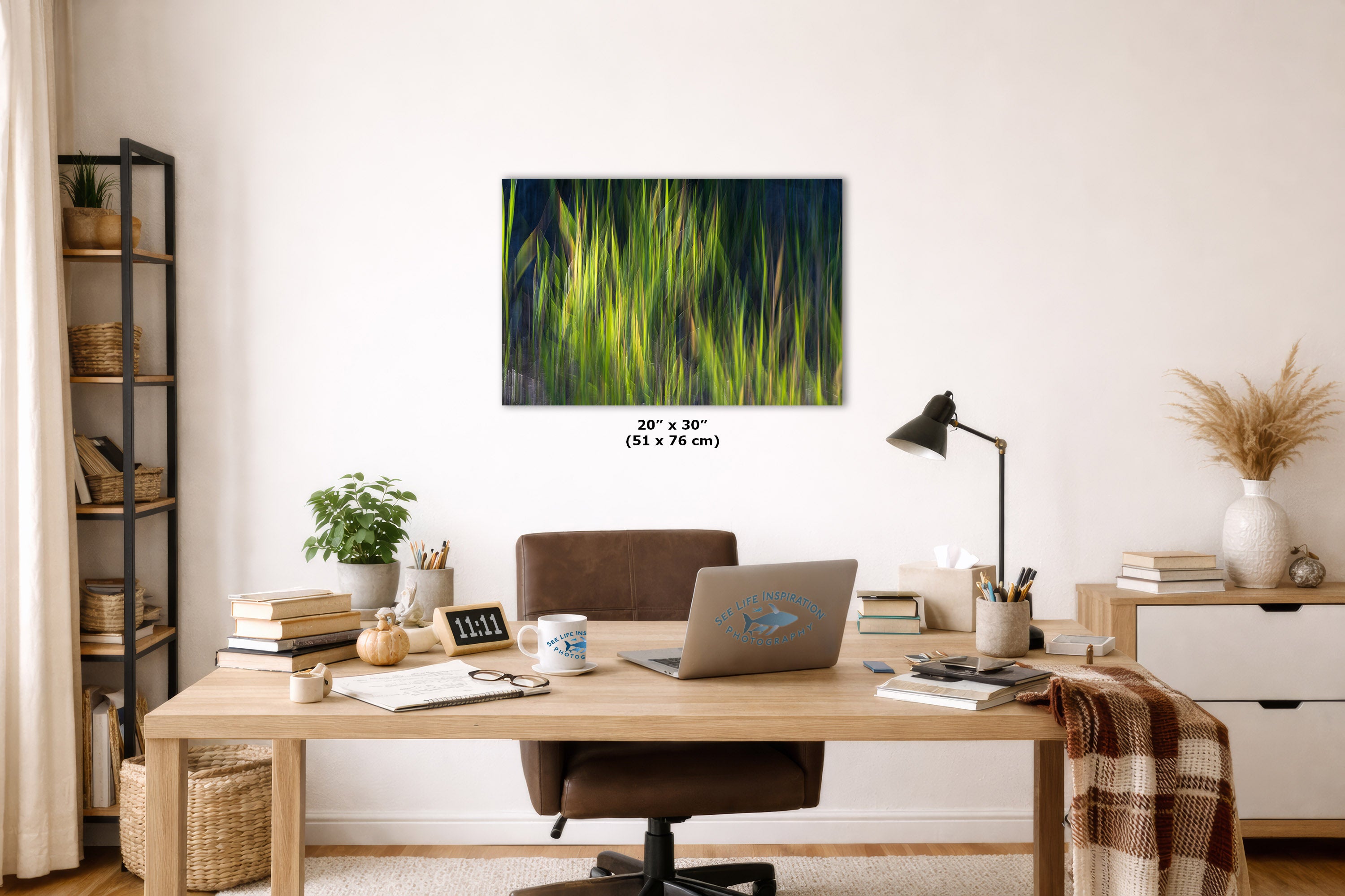 A framed wall art print depicting vibrant green grass against a dark background, styled in a modern home office setting above a wooden desk with a laptop, lamp, and shelves.