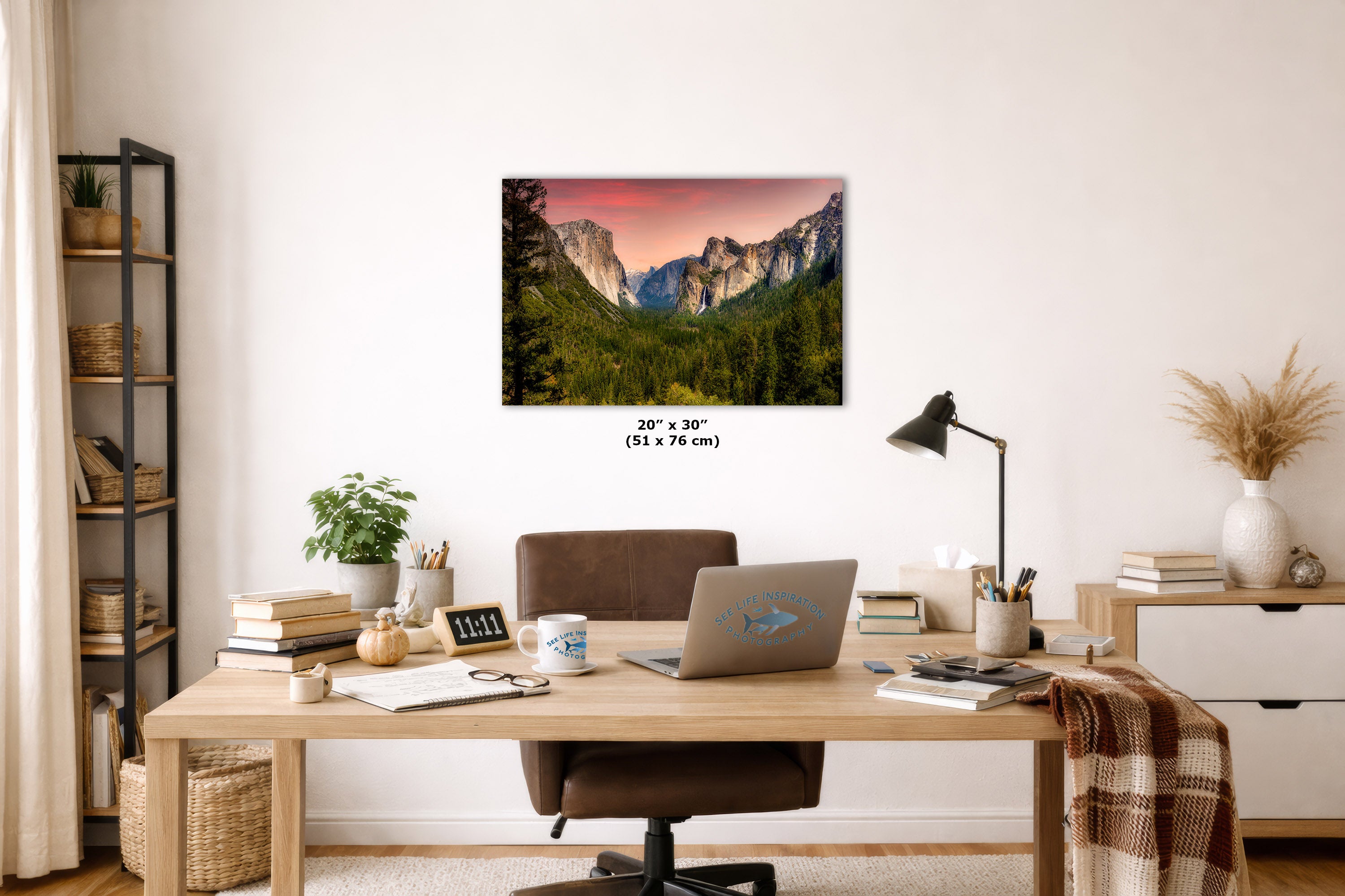 A wall art print showing a mountain valley landscape with green trees and a pink-orange sky, displayed above a wooden desk in a styled home office. The print includes size text reading 20 x 30 inches (51 x 76 cm).
