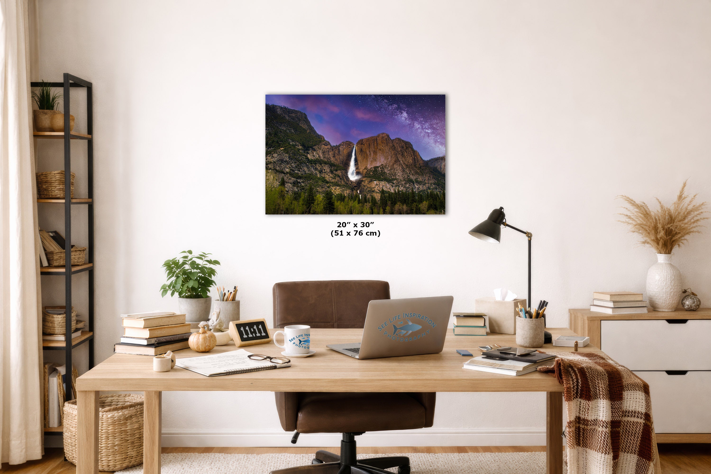 A wall art print showing a waterfall in a mountain landscape under a purple sky, displayed above a wooden desk in a styled home office setting.