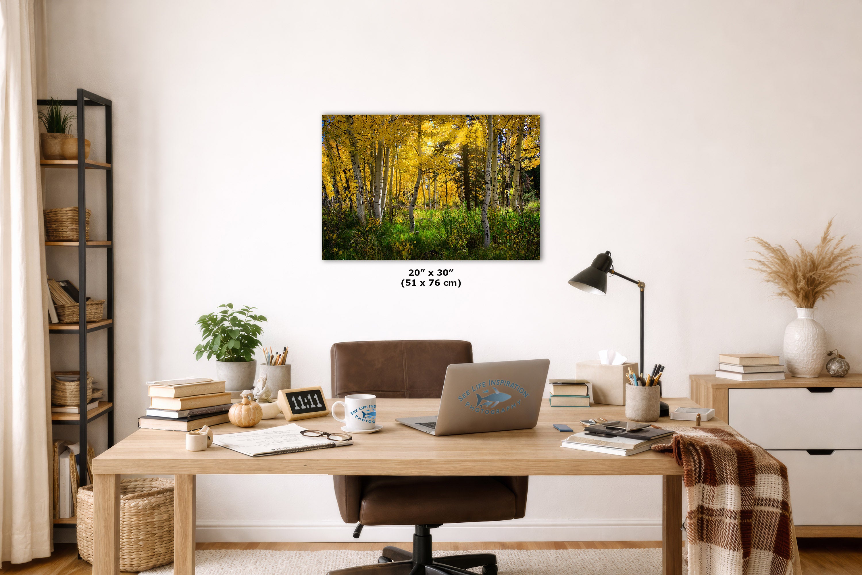 A wall art print showing a golden yellow autumn forest hangs on a white wall above a wooden desk in a styled home office setting.