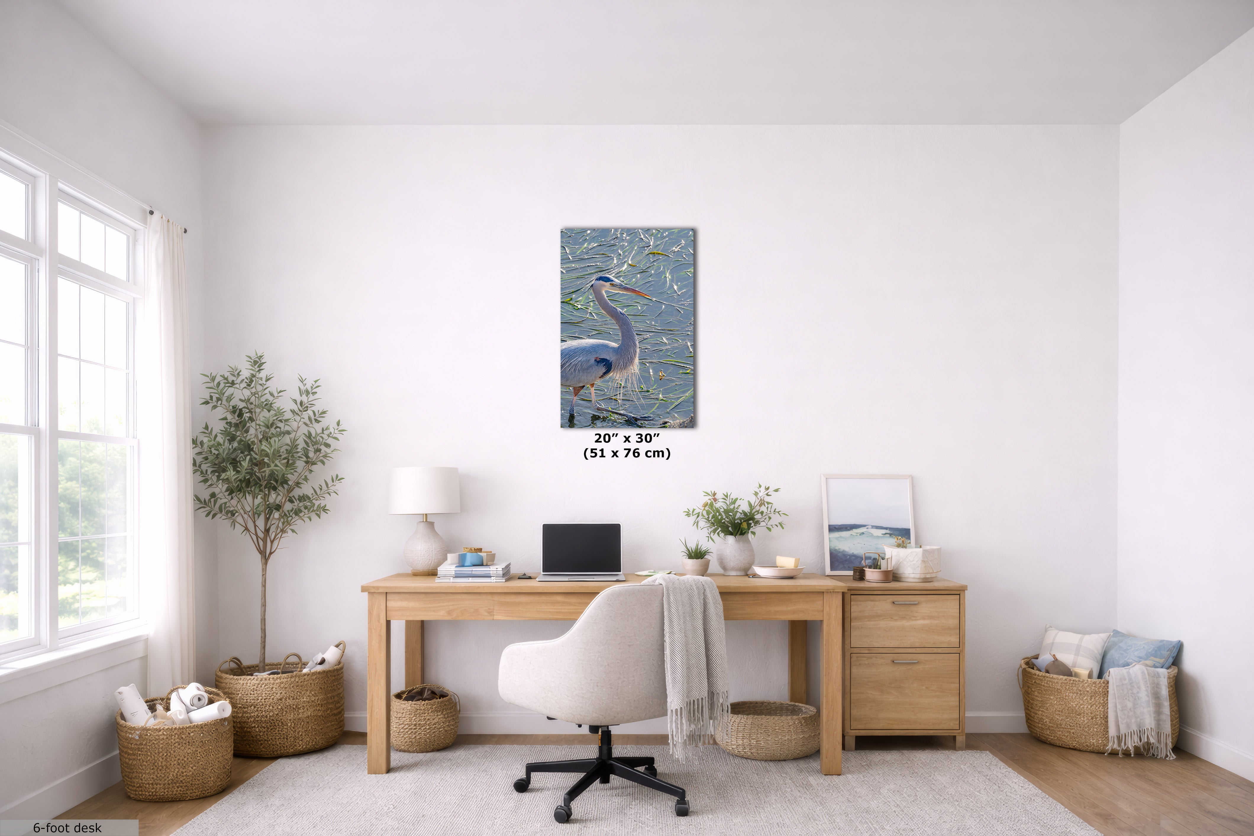 A framed vertical canvas print of a blue heron over water, displayed on a white wall in a bright, modern home office setting above a wooden desk.