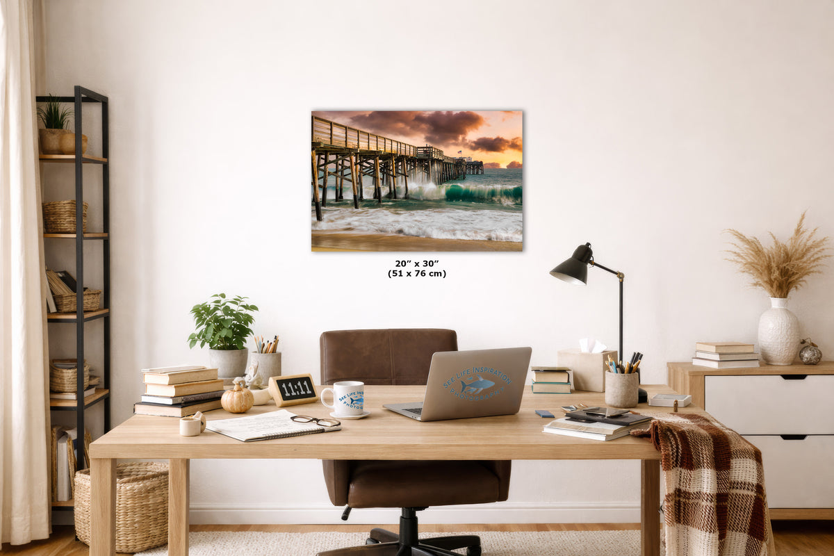 A wall art print depicting a pier over ocean waves at sunset, displayed above a wooden desk in a styled home office. The print includes size text reading 20" x 30" (51 x 76 cm).