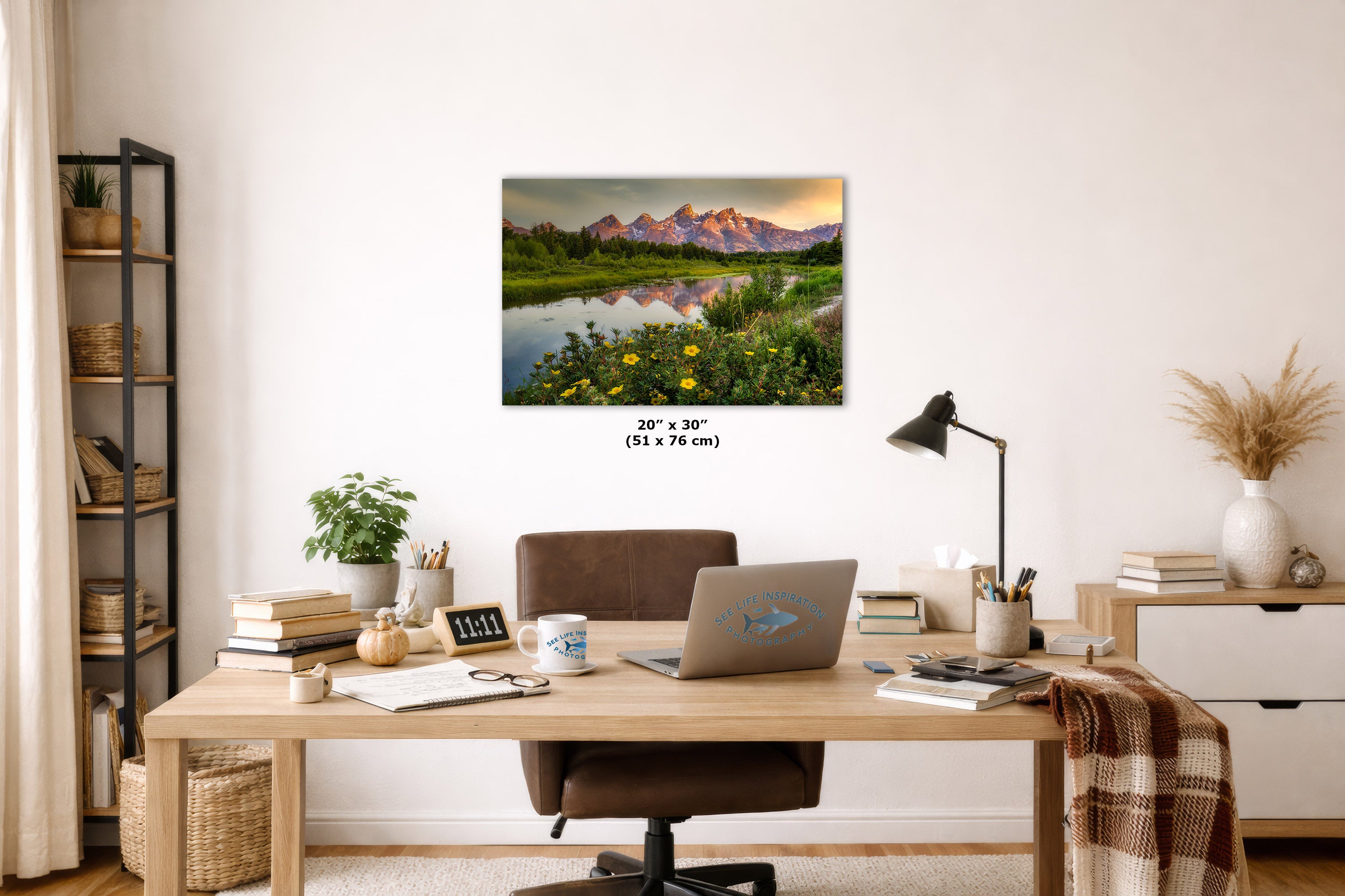 A wall art print showing a mountain landscape reflected in a calm lake with wildflowers, displayed above a wooden desk in a styled home office. The print includes size text reading 20 x 30 inches (51 x 76 cm).