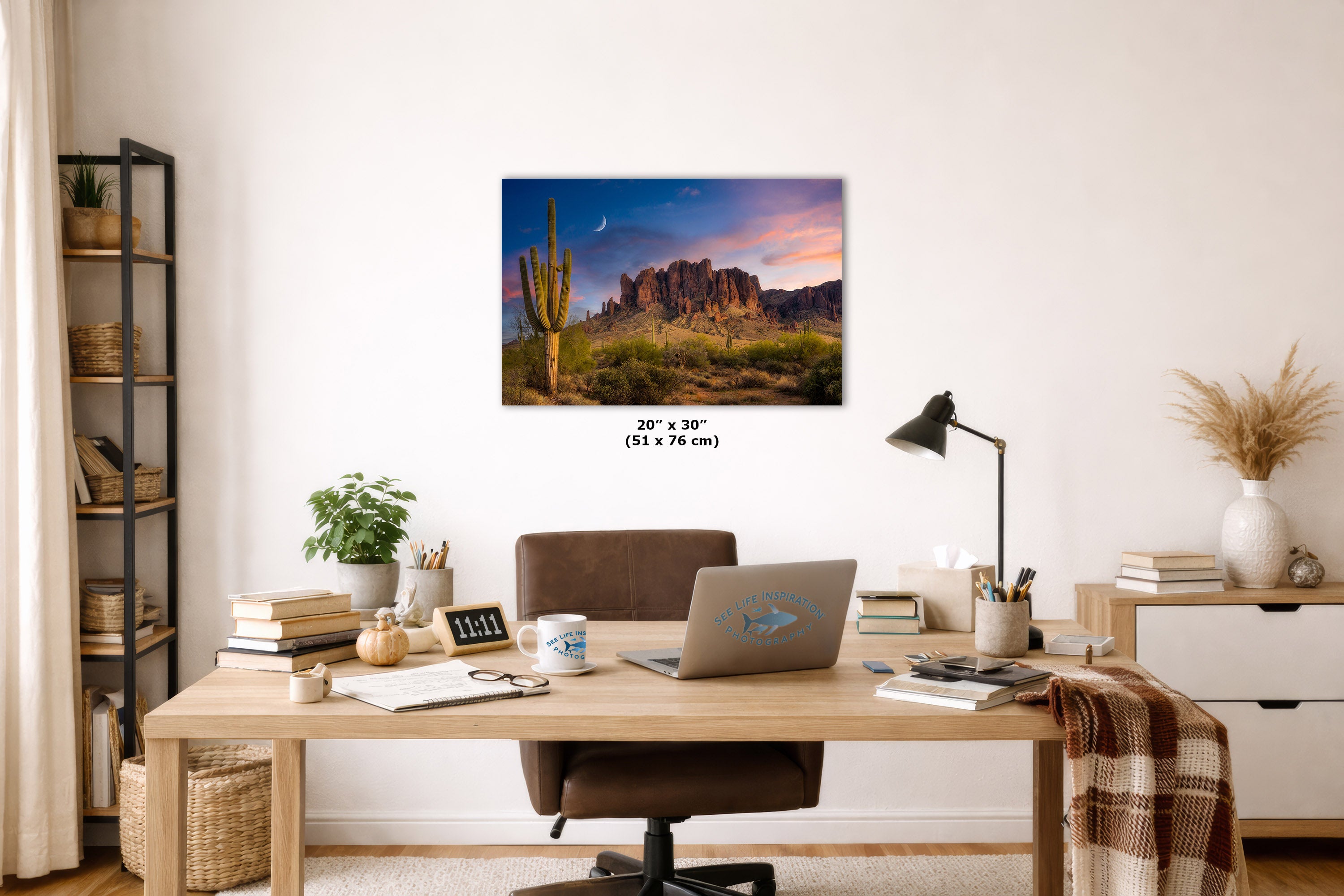A wall art print showing a desert landscape with a saguaro cactus and rock formations under a colorful sunset sky, displayed above a wooden desk in a styled home office setting.