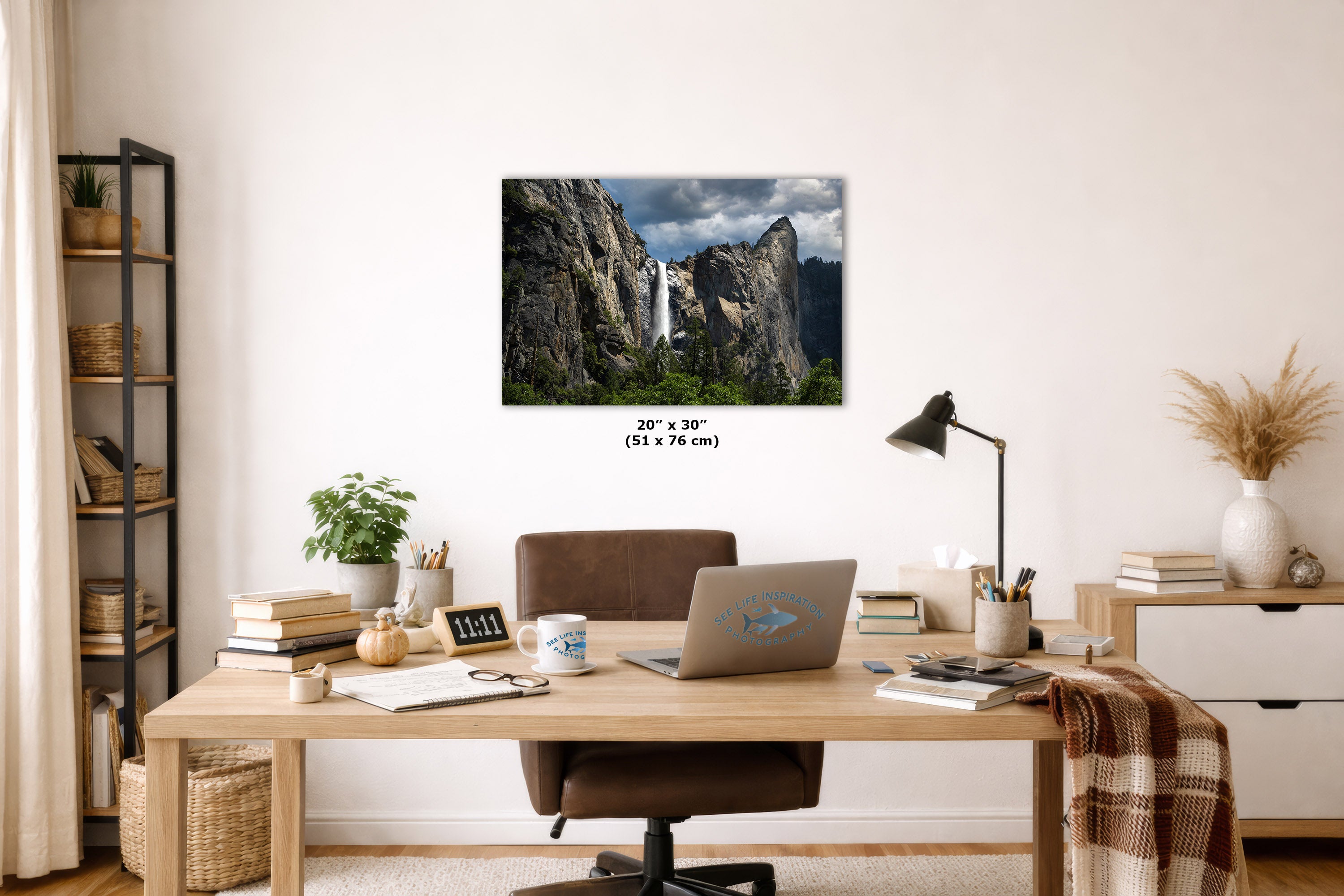 A wall art print showing a waterfall between rocky cliffs hangs above a wooden desk in a styled home office. The print includes size text reading 20 x 30 inches (51 x 76 cm).