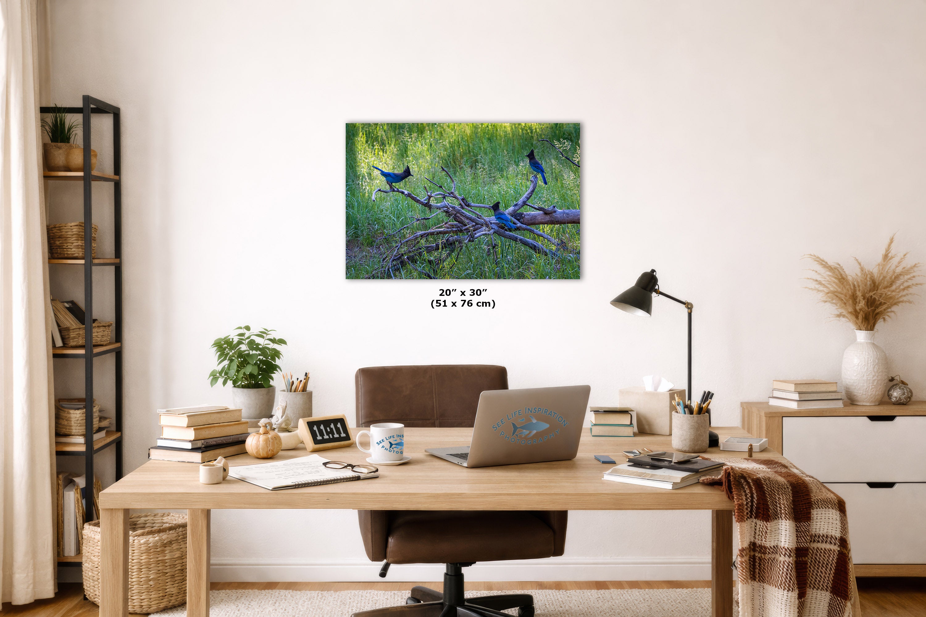 A wall art print showing blue birds perched on branches in a grassy green setting, displayed above a wooden desk in a styled home office. The print includes size text reading 24 x 36 inches (51 x 76 cm).