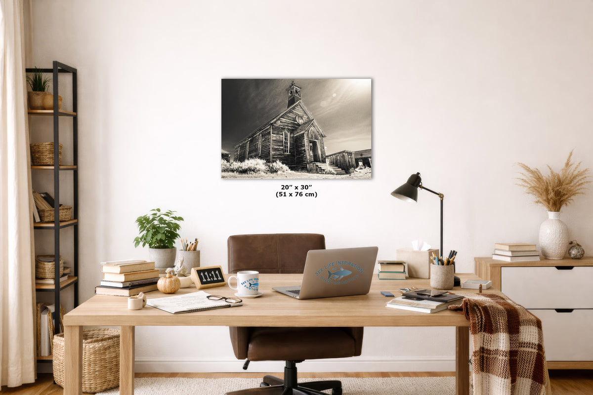 An art print of a rustic old church building in snow, displayed on a white wall in a styled home office setting above a wooden desk with a laptop and chair.