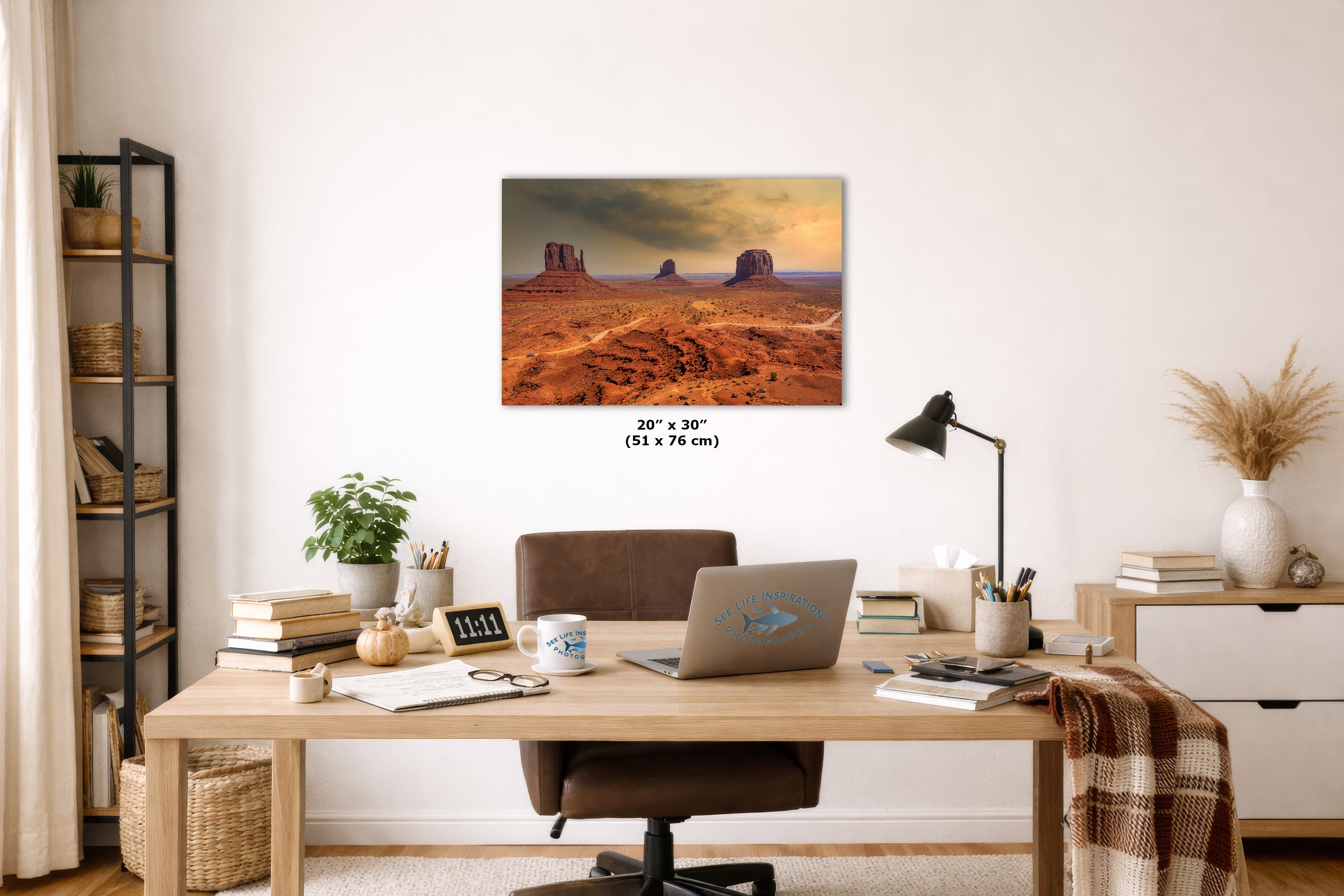 A wall art print depicting a desert landscape with rock formations under a warm sky, displayed above a wooden desk in a styled home office setting. The print includes size text reading 20 x 30 cm.