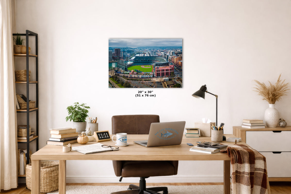 A wall art print showing an aerial view of a baseball stadium and surrounding cityscape, displayed above a wooden desk in a styled home office. The print includes size text reading 20 x 30 inches.
