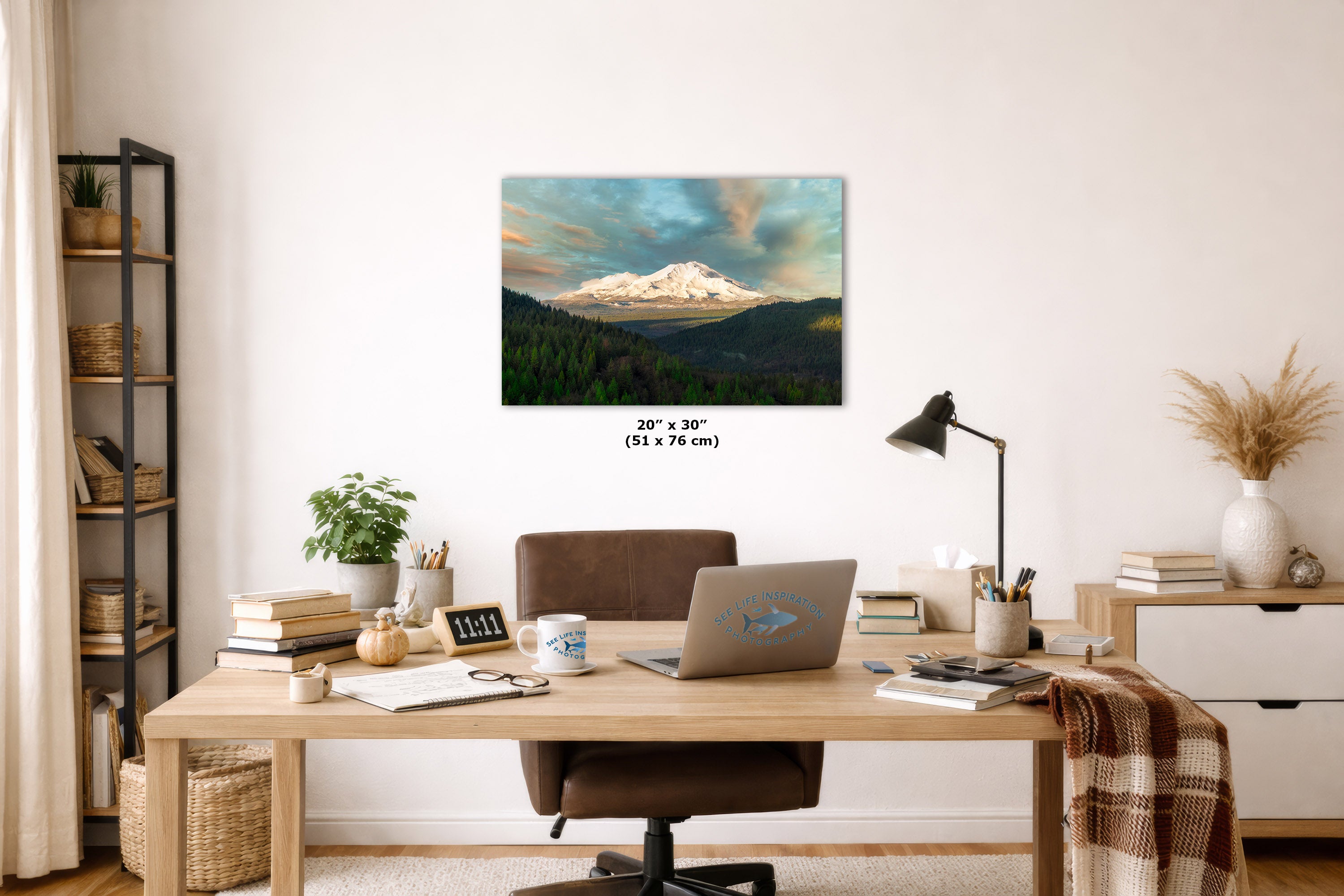 A wall art print depicting a snow-capped mountain under a dramatic cloudy sky, displayed above a wooden desk in a styled home office setting. The print includes size text indicating 20 x  30 inches.