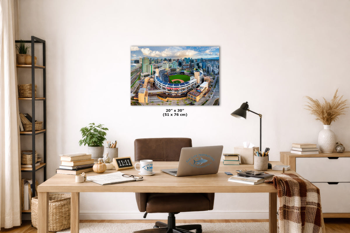 A wall art print of a stadium cityscape hangs above a wooden desk in a styled home office. The print shows a blue stadium surrounded by a city skyline under a cloudy sky.