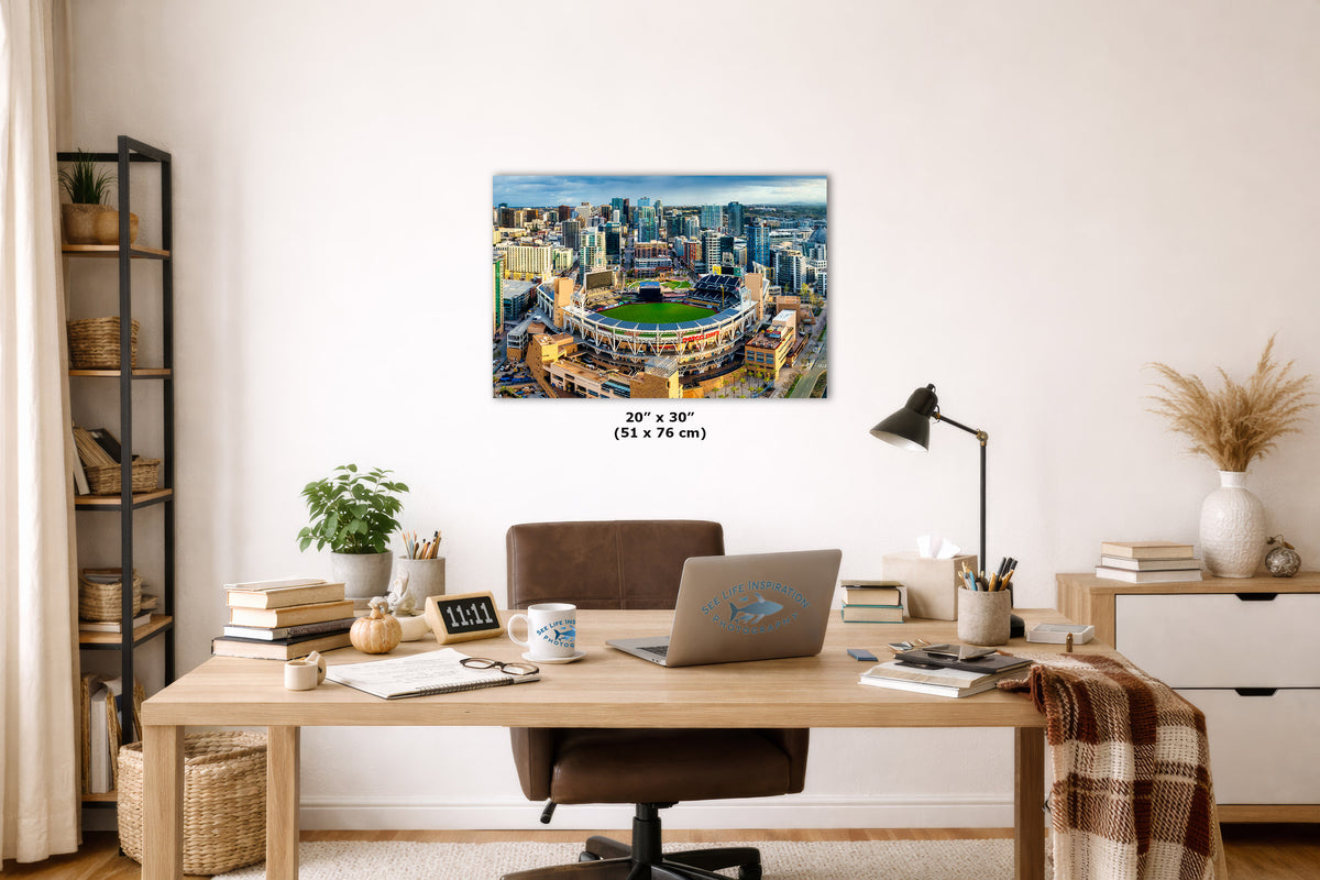 A wall art print of a stadium cityscape hangs above a wooden desk in a styled home office. The print shows a detailed aerial view of a sports stadium surrounded by a dense urban skyline.