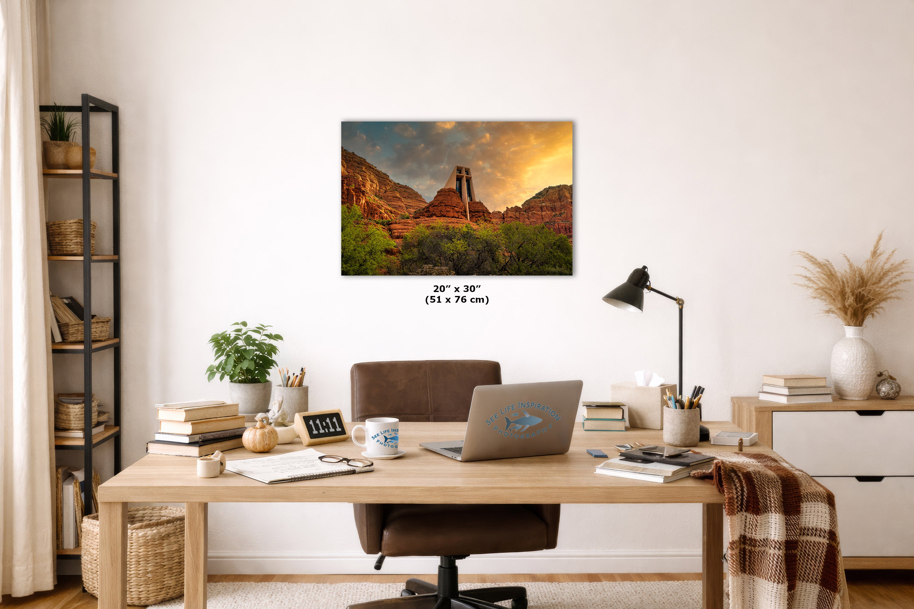 A wall art print depicting a desert canyon landscape with a tower under a dramatic sunset sky, displayed above a wooden desk in a styled home office setting. The print includes size text reading 20 x 76 inches.