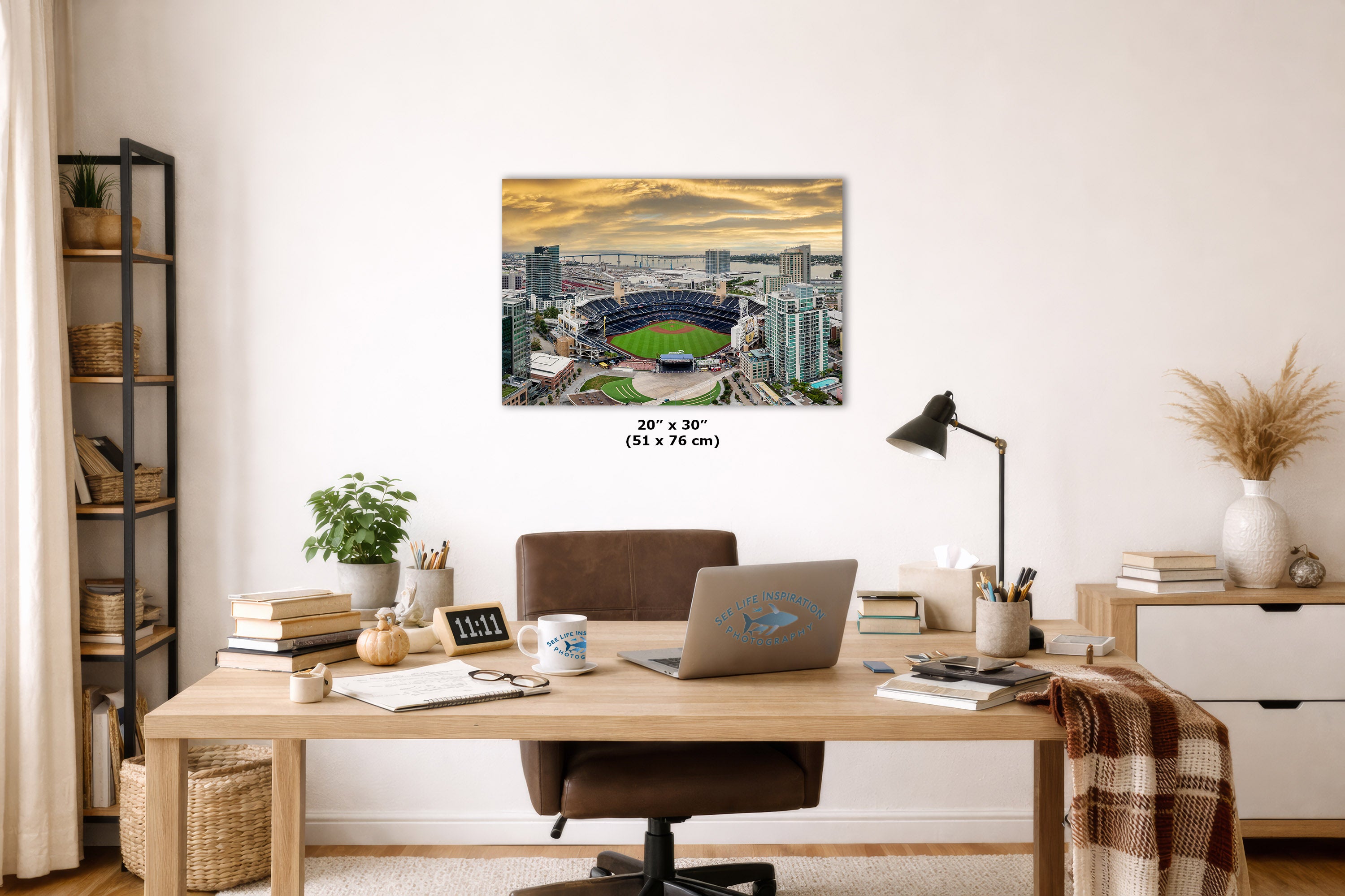 A rectangular wall art print showing an aerial stadium scene at sunset, displayed above a wooden desk in a styled home office. The print includes size text reading 20 x 30" (51 x 76 cm).