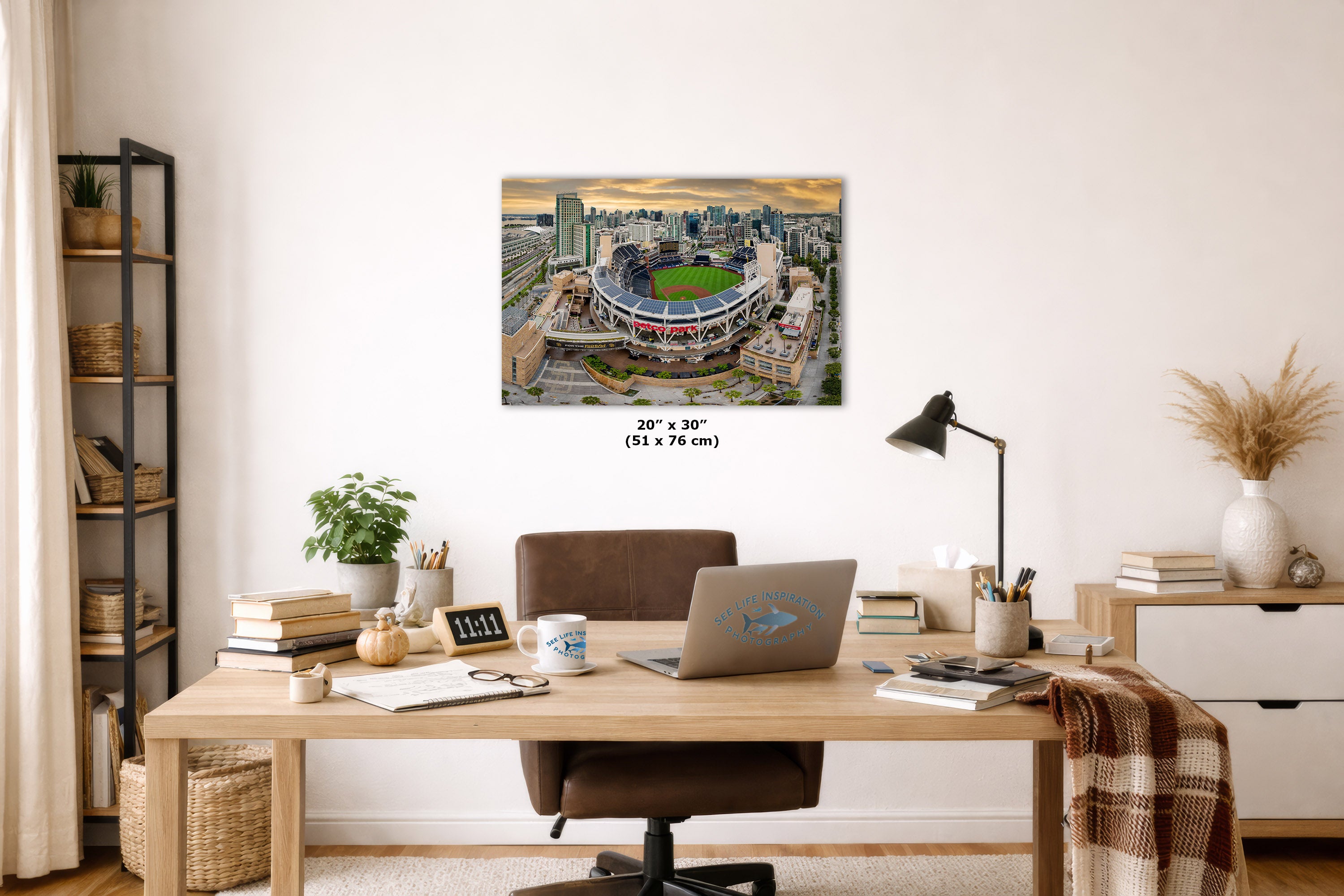 A wall art print showing an aerial view of a baseball stadium and surrounding city skyline, displayed above a wooden desk in a styled home office. The print includes size text reading 20 x 30 inches.