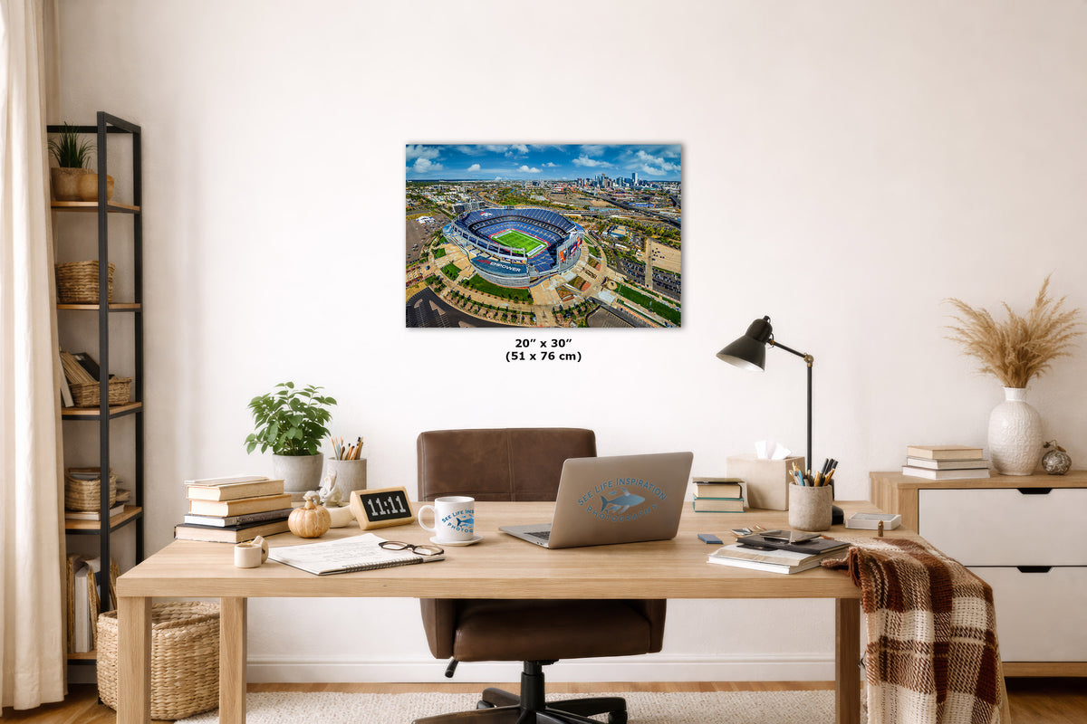 A wall art print showing an aerial view of a blue stadium under a cloudy sky, displayed above a wooden desk in a styled home office. The print includes size text reading 20 x 30 inches (51 x 76 cm).